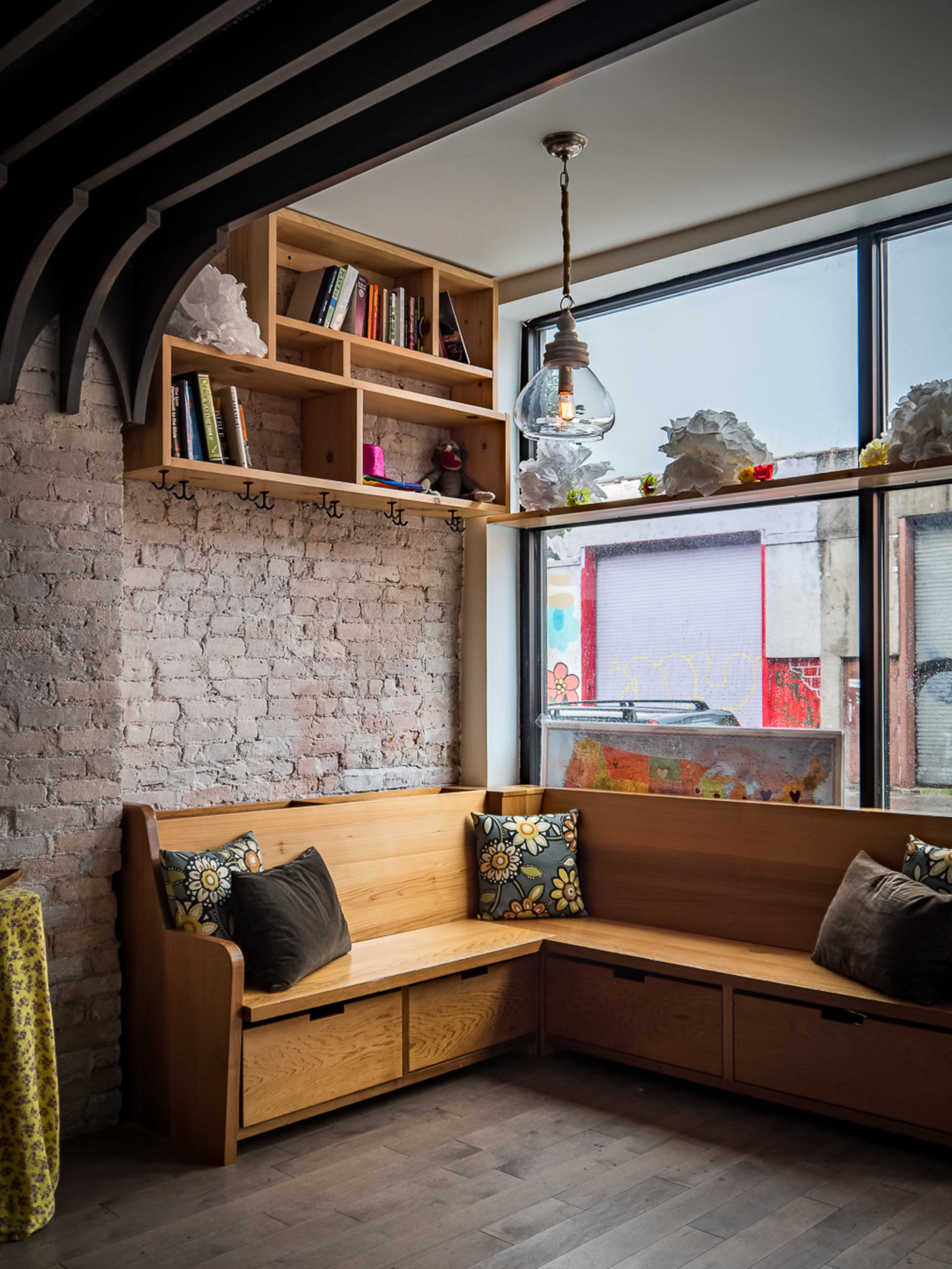 The image shows a cozy corner seating area with built-in wooden benches, shelves filled with books, and large windows providing natural light.