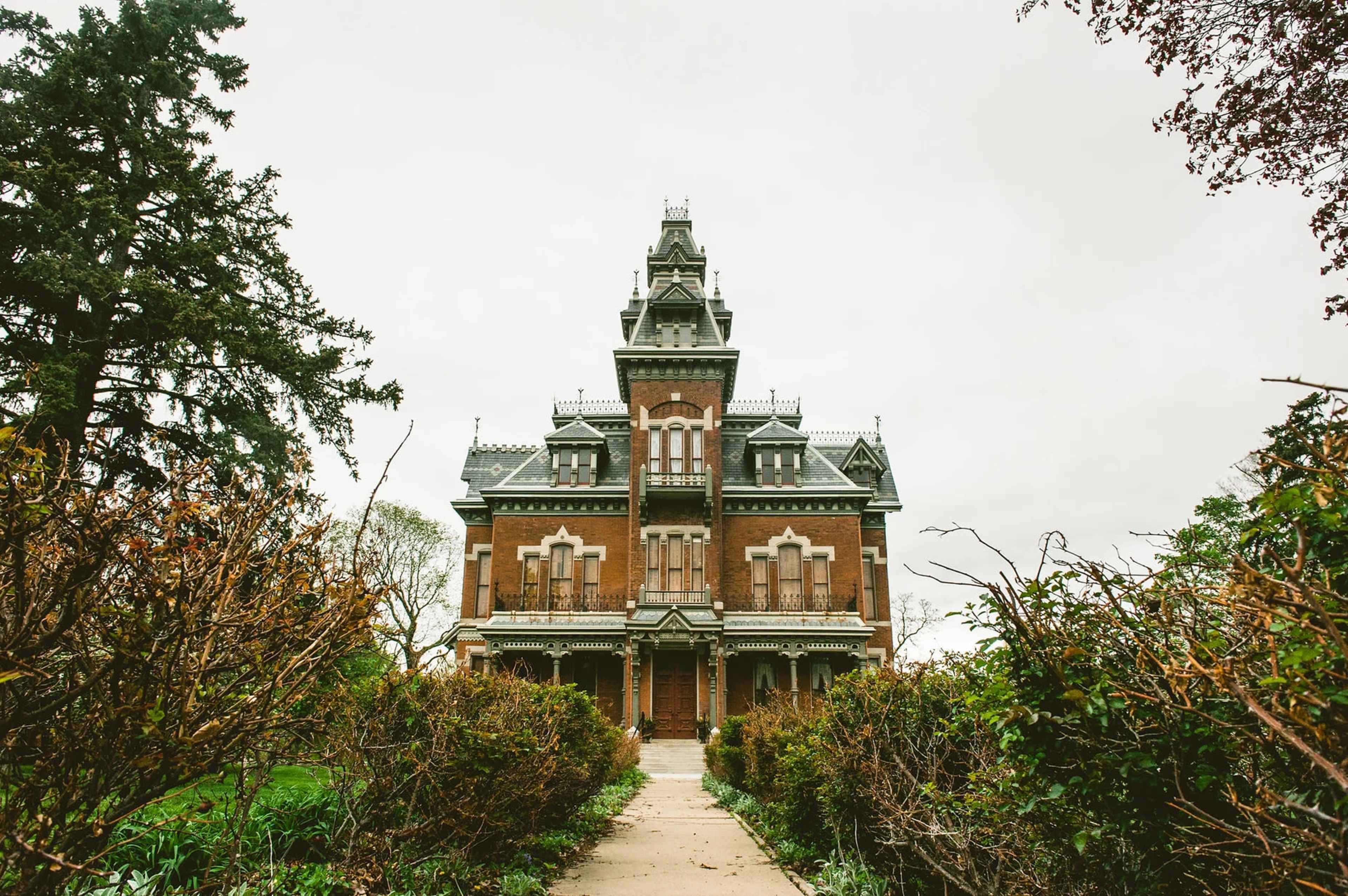 A large, two-story Victorian mansion with a pointed turret and ornate details stands at the end of a pathway, surrounded by overgrown shrubs.