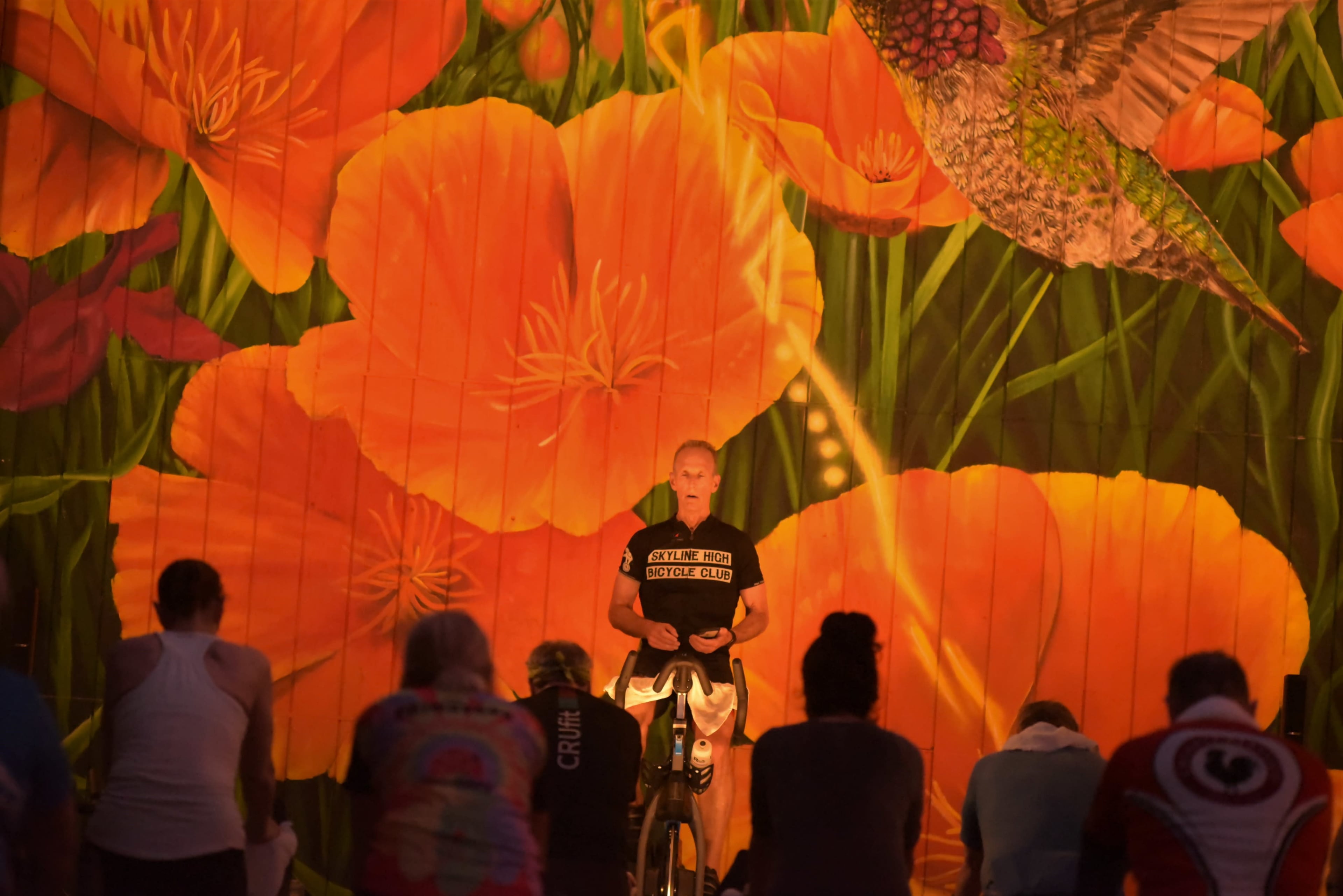 A fitness instructor leads a cycling class in front of a large mural of orange flowers and a hummingbird.