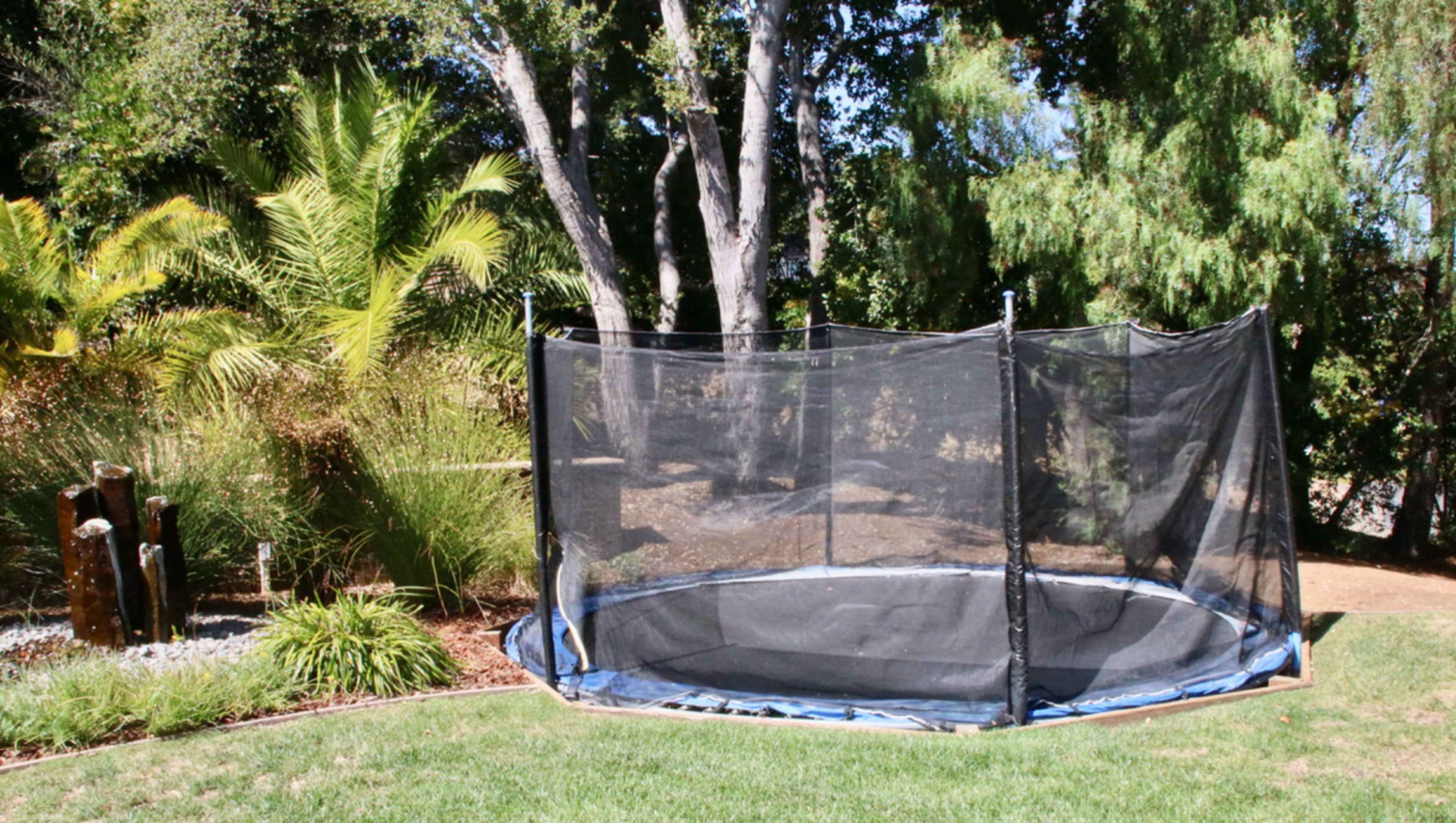 A black trampoline enclosed by a safety net is positioned on a grassy area surrounded by trees and foliage.