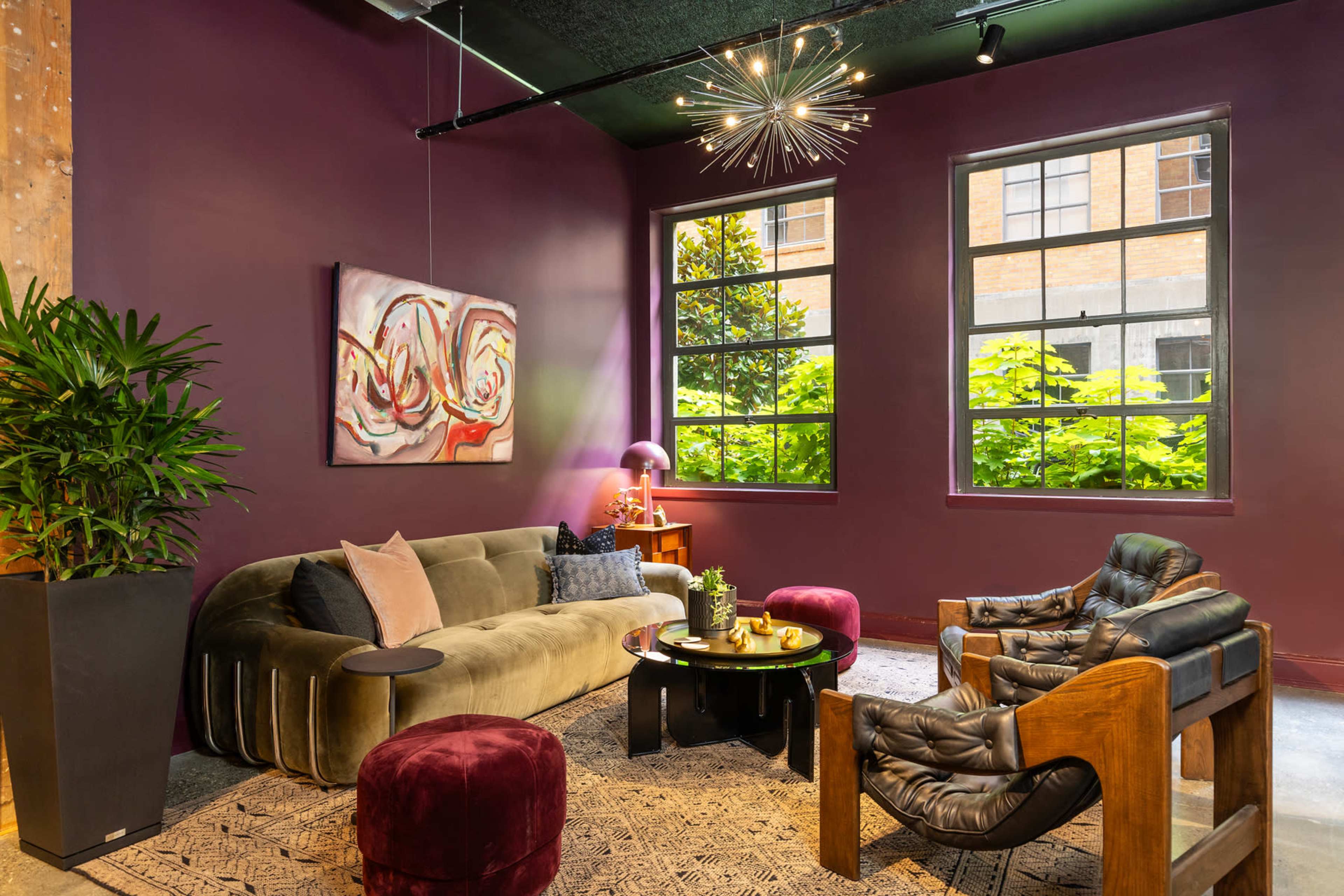 A modern lounge features a velvet sofa, leather chairs, and a decorative art piece against a purple wall, with large windows allowing natural light and views of greenery.