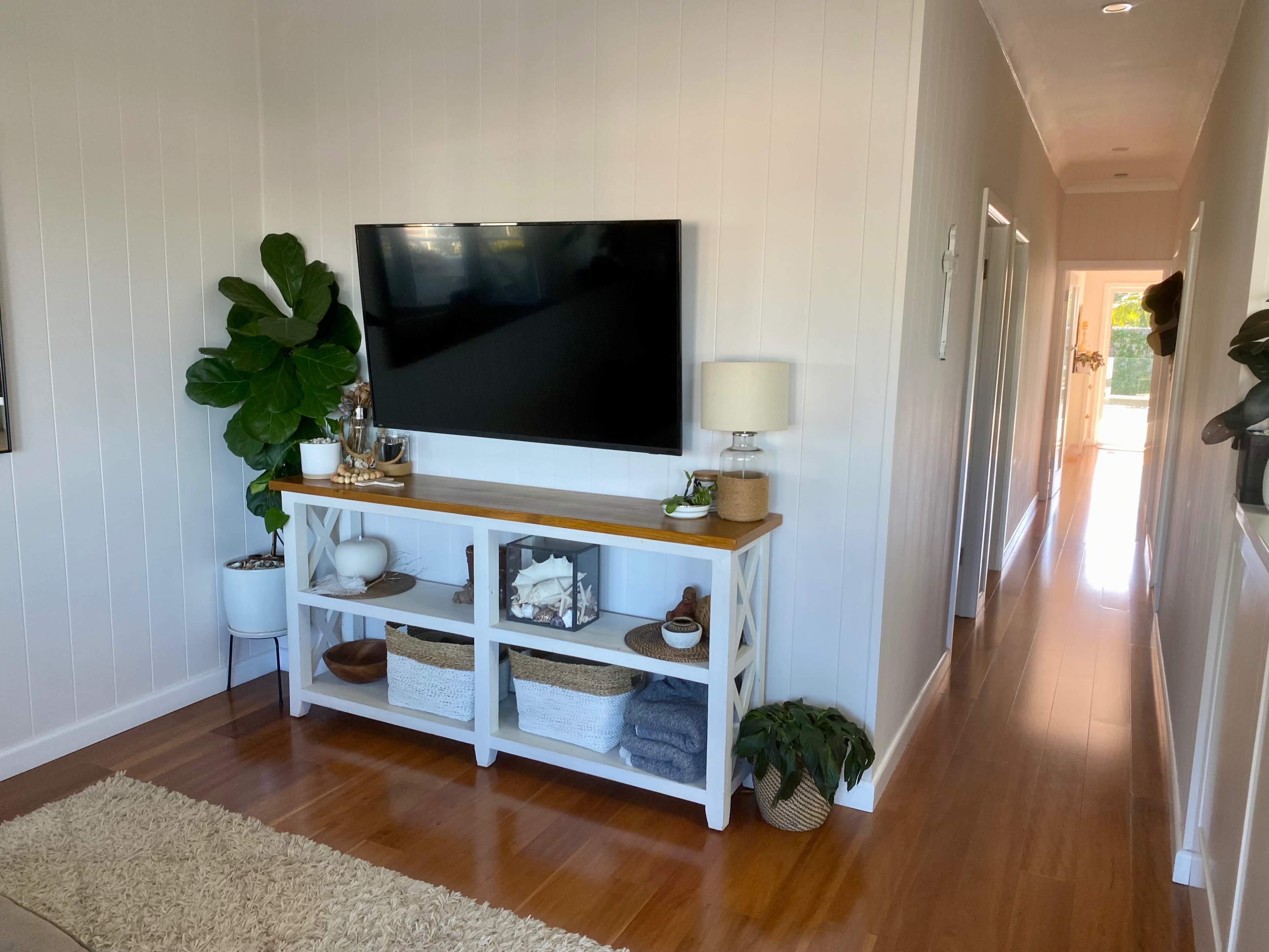 A hallway that features a television mounted on the wall and a white console table with decorative items and storage baskets, alongside a potted plant.