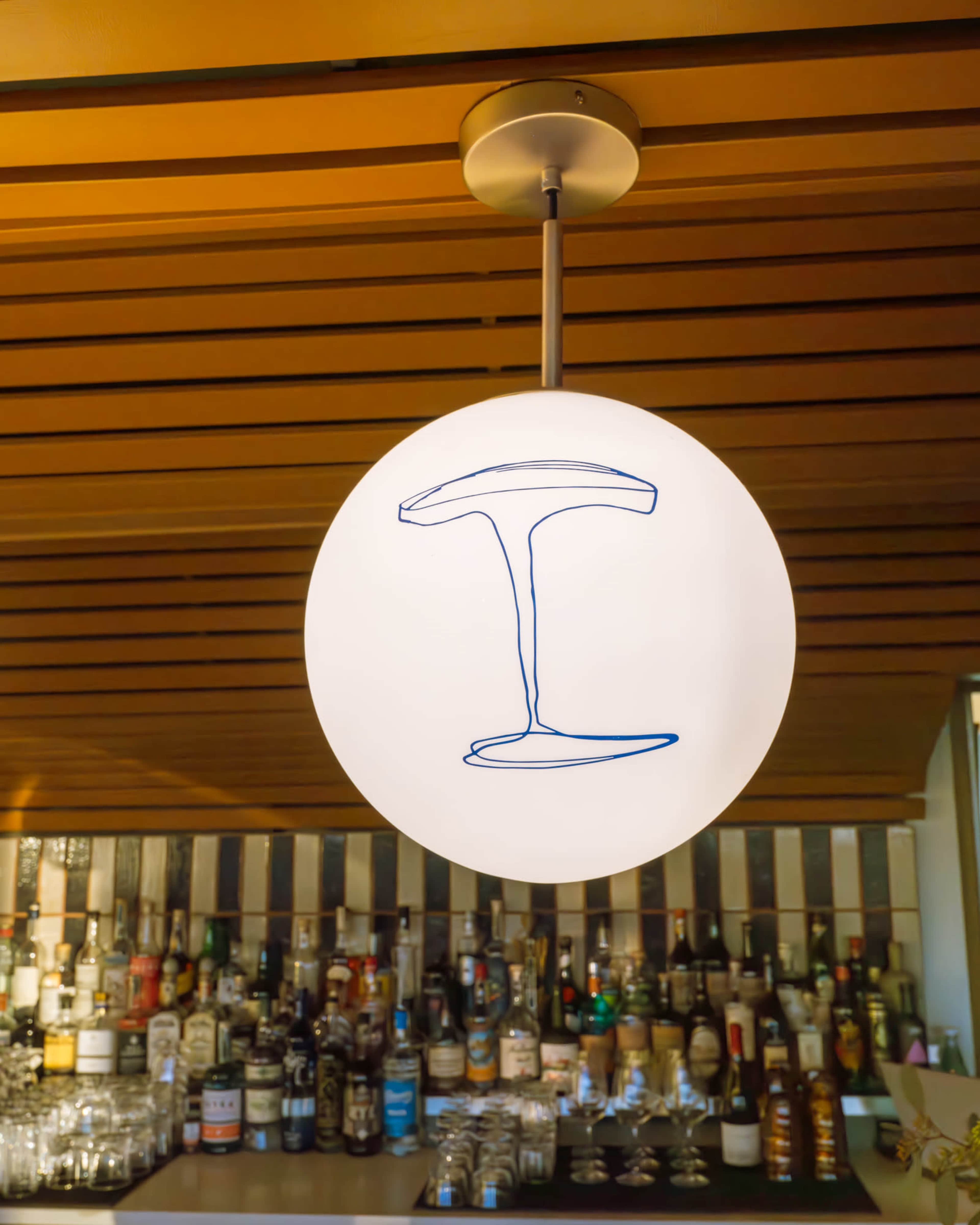 A round pendant light featuring a graphic drawing of a martini glass hangs above a bar stocked with various bottles.