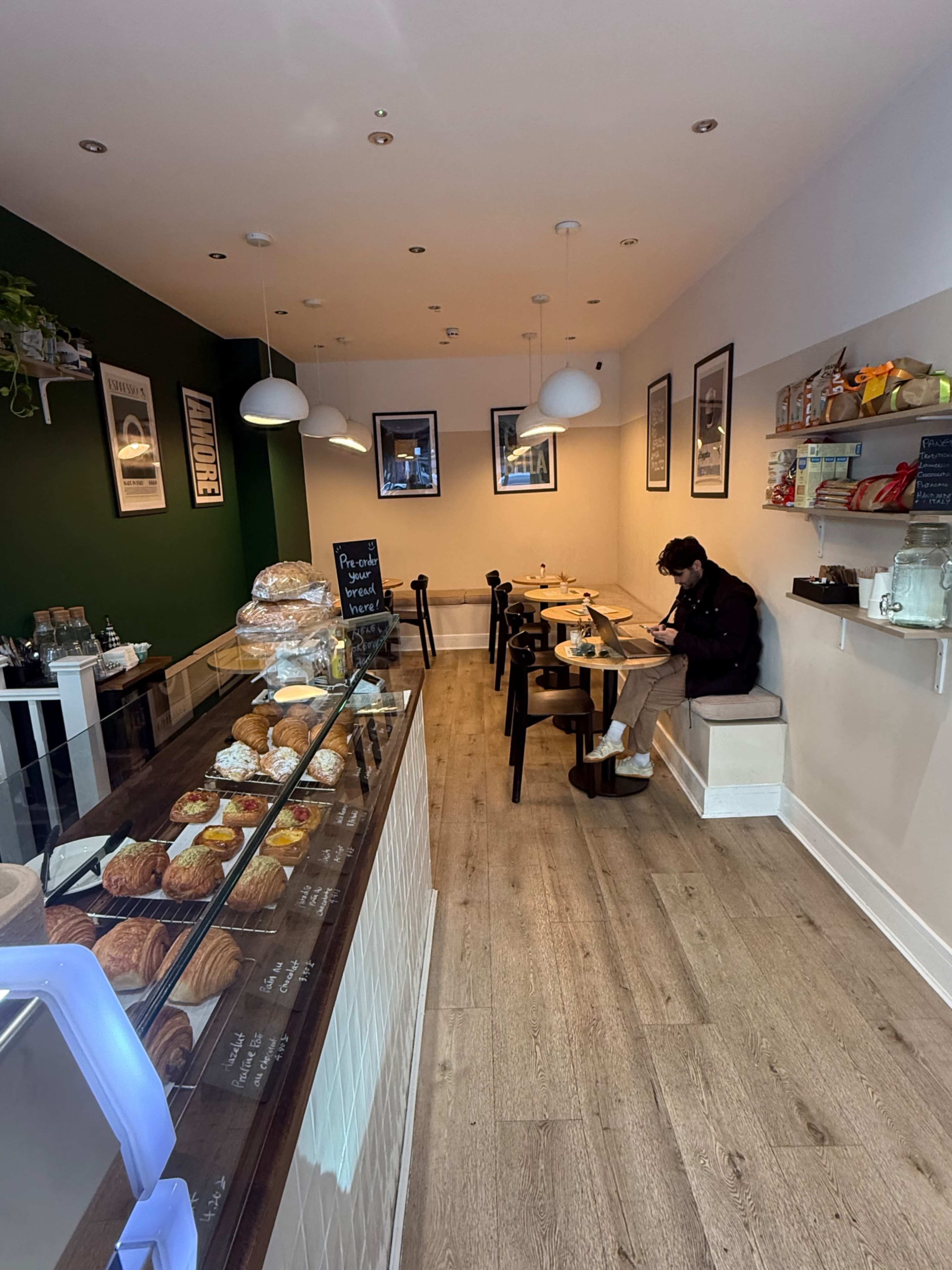 The image shows a small café with a display case of baked goods on the left and a person sitting at a table on the right.
