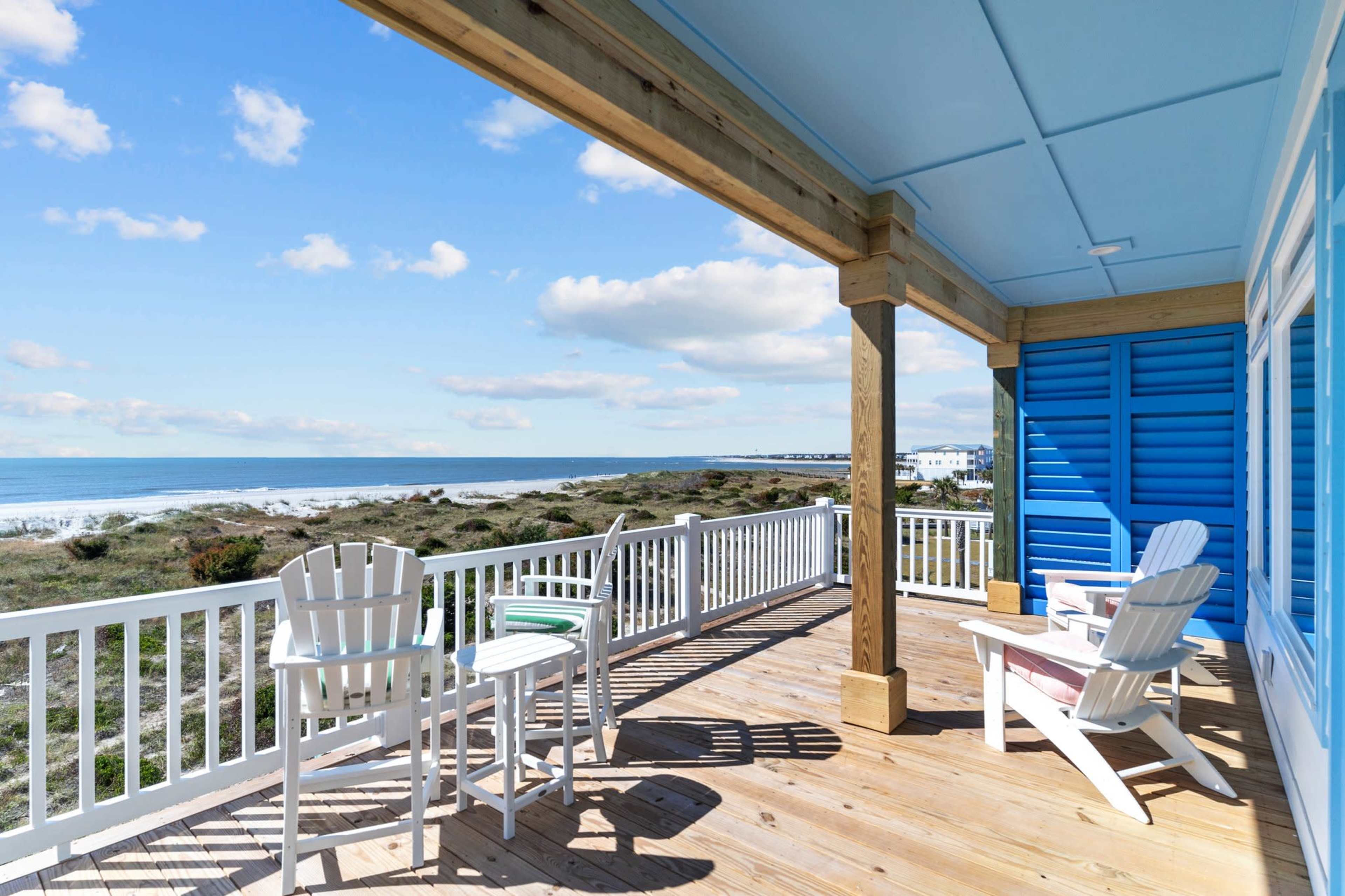 A wooden deck with white adirondack chairs overlooking a beach and ocean under a blue sky.