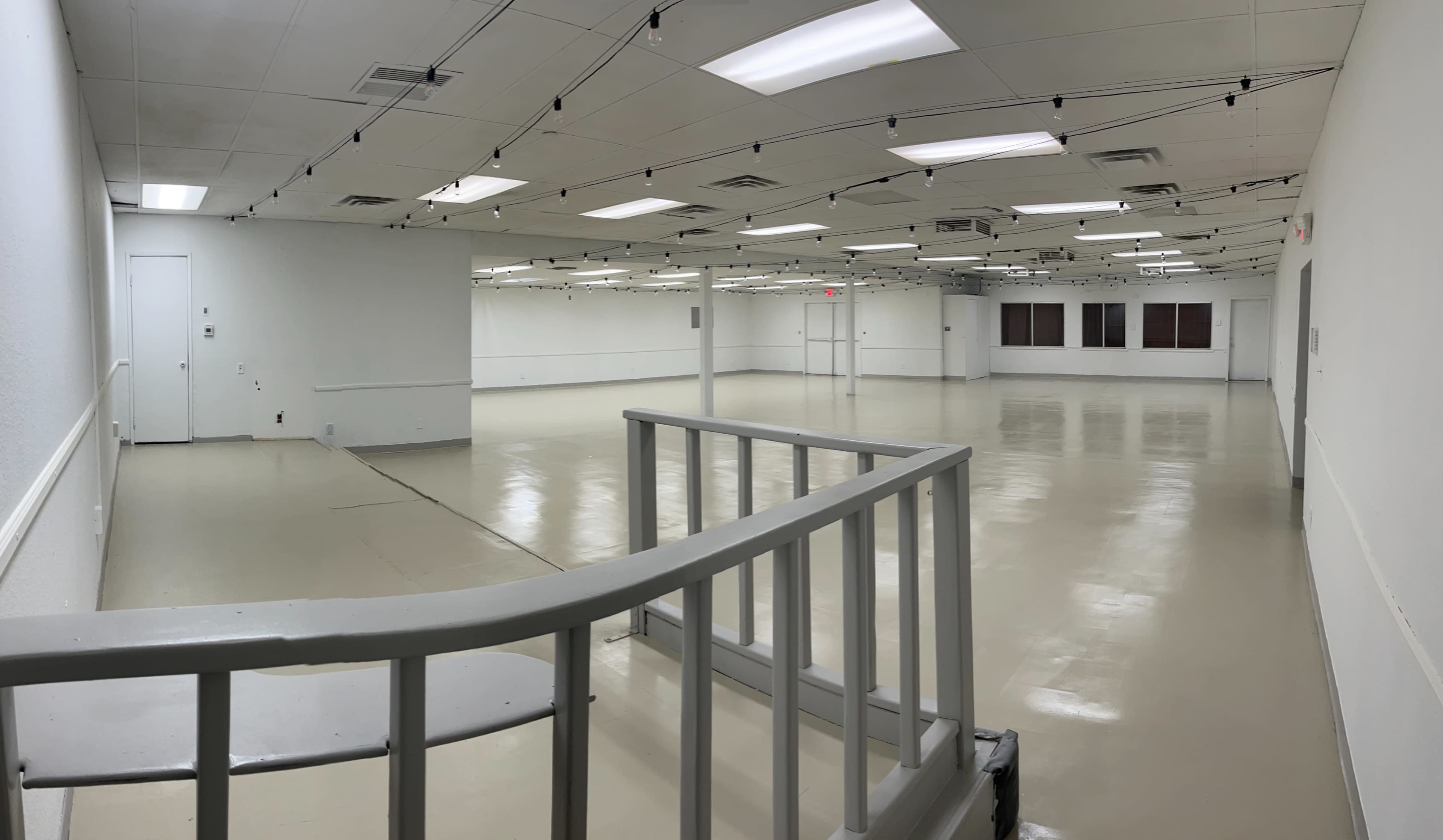 An empty, well-lit room with a glossy floor and white walls, featuring overhead lights and a railing in the foreground.