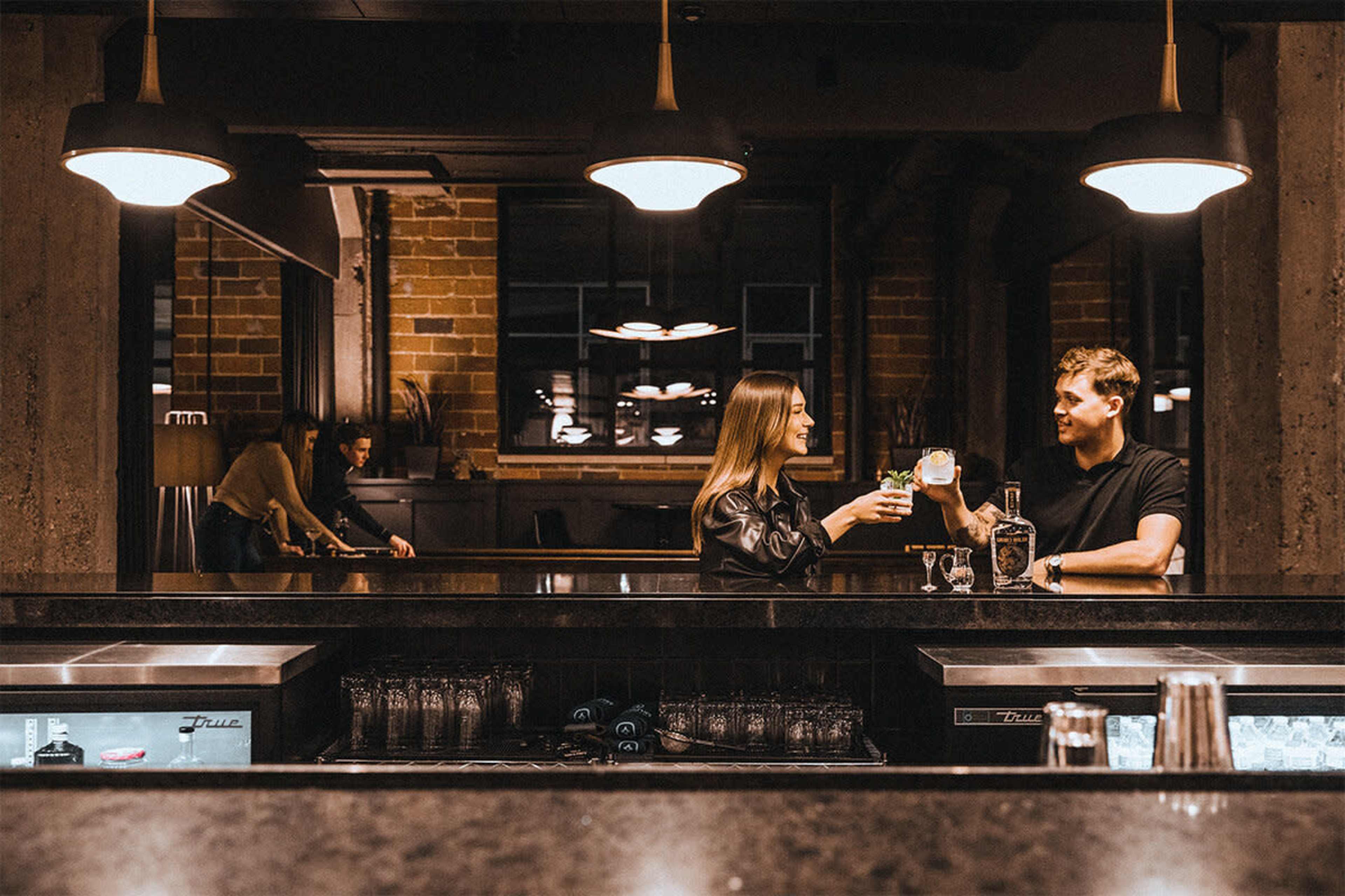 Two people are clinking glasses at a dimly lit bar while another person is preparing drinks in the background.
