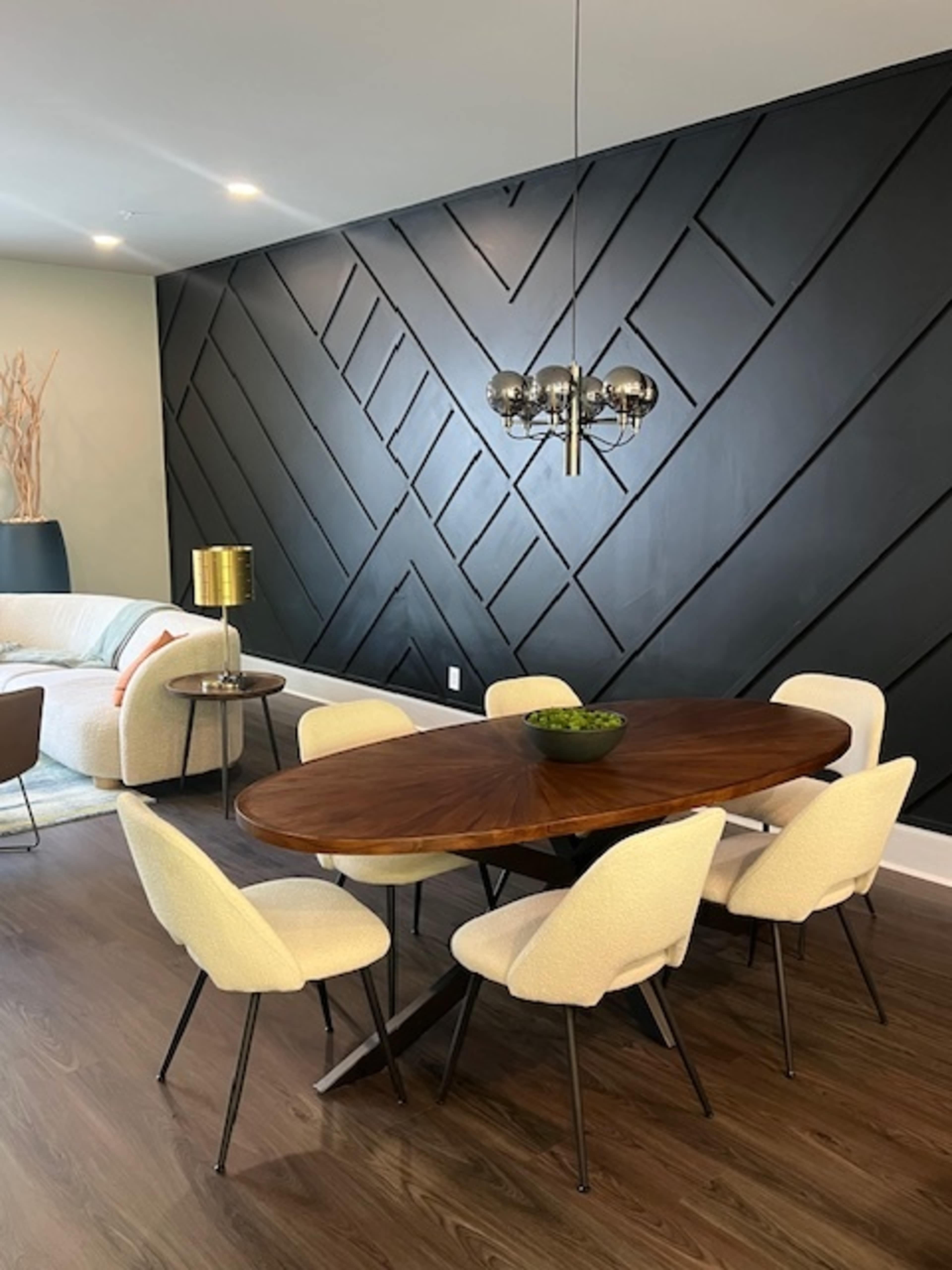 The image features a modern dining area with a large oval wooden table surrounded by six light-colored upholstered chairs, set against a wall with a geometric black design.