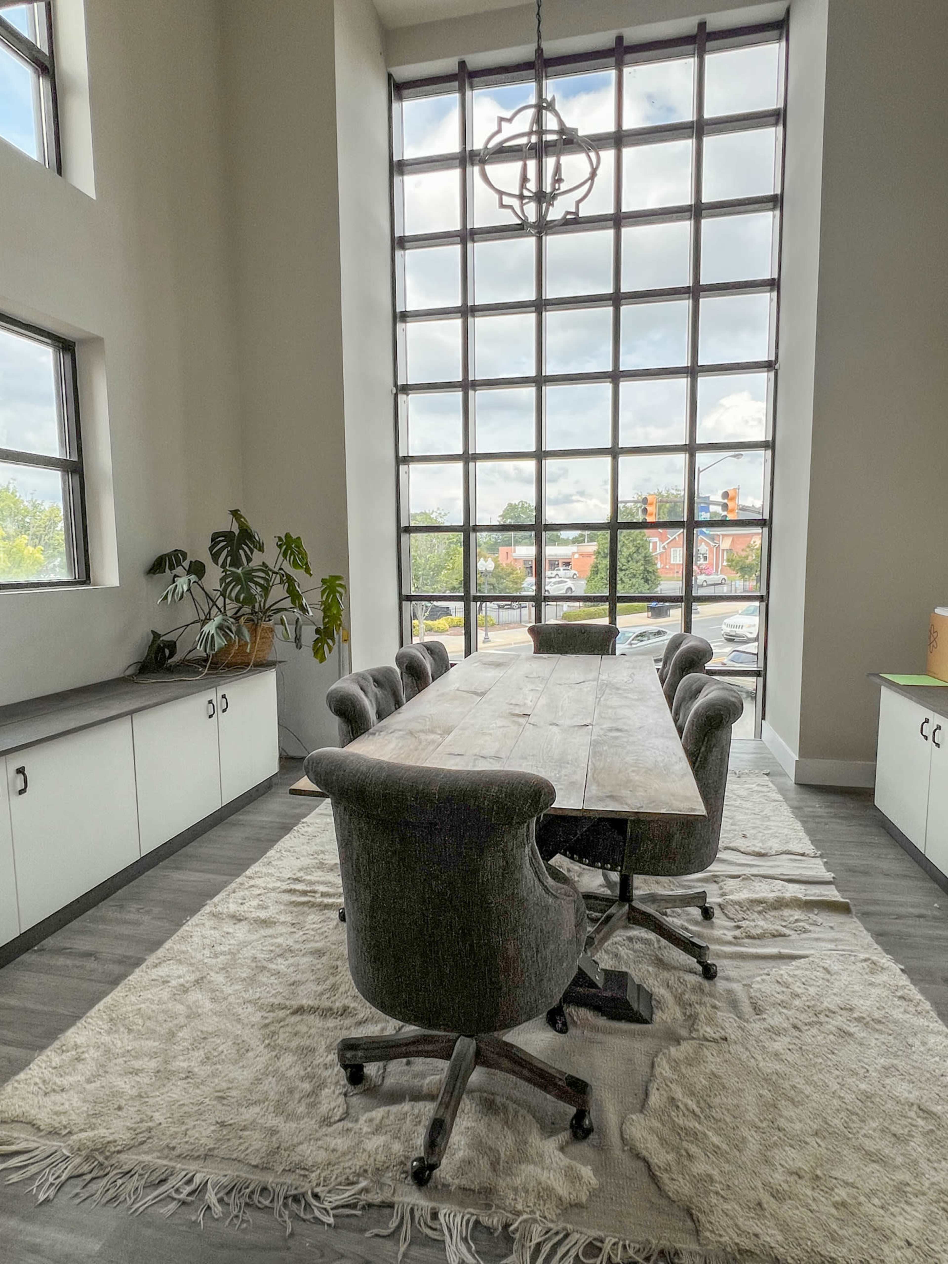 A long wooden conference table with upholstered chairs is placed in a well-lit room featuring large windows and a potted plant.