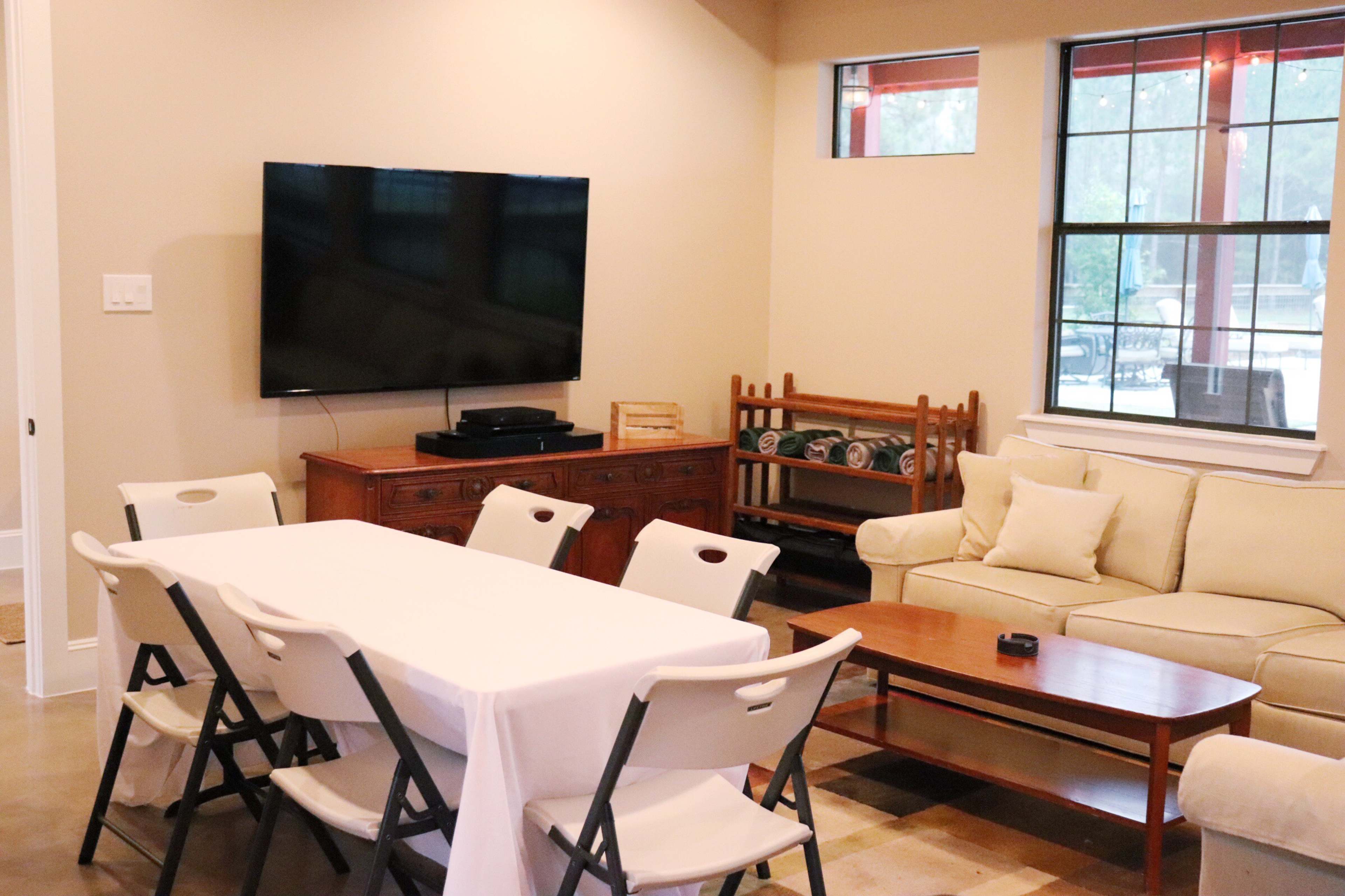 The image shows a living area with a television mounted on the wall, a wooden sideboard, a coffee table, a couch, and a dining table set with folding chairs.