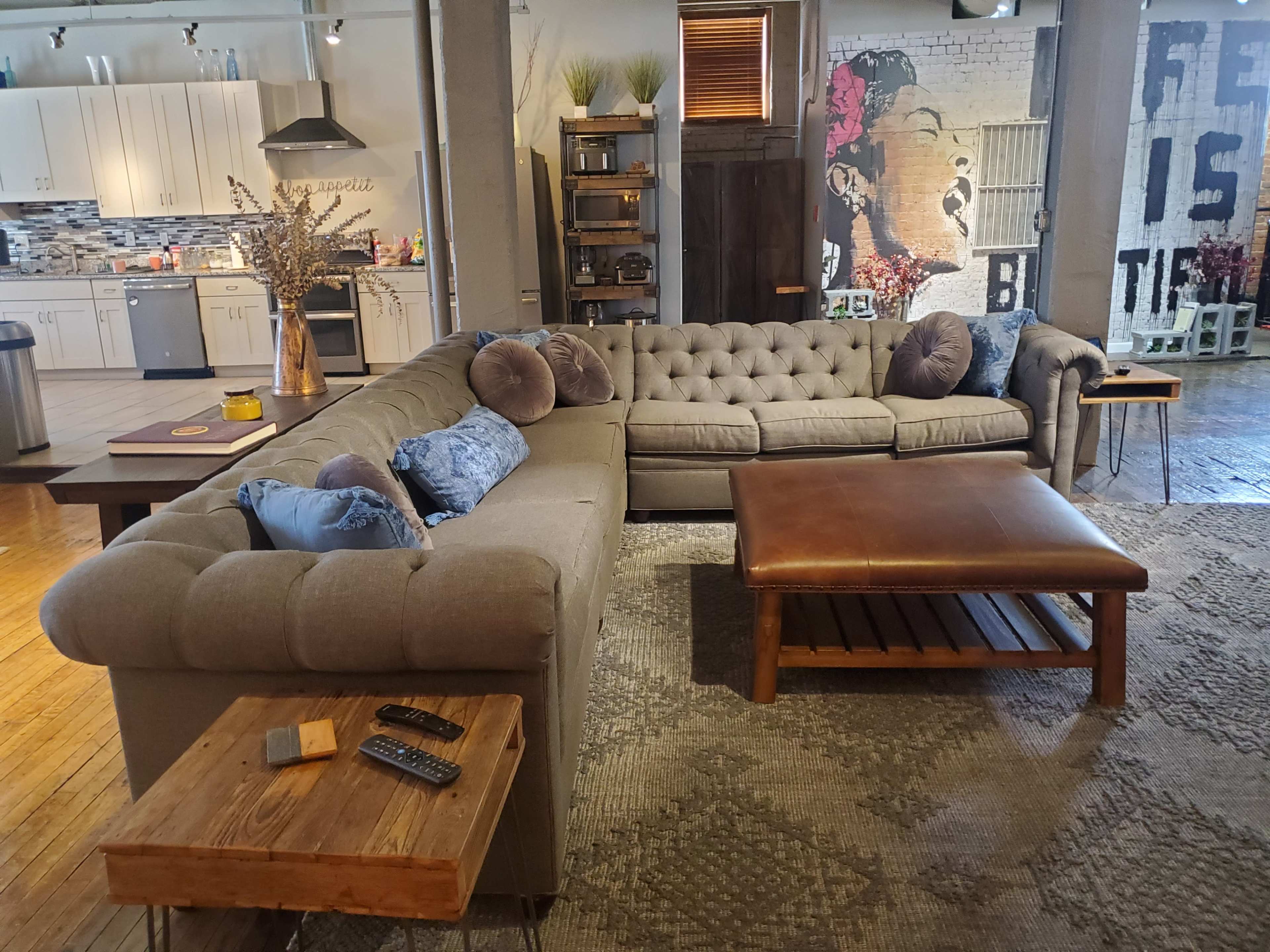 The living area features a spacious tufted sectional sofa with decorative pillows, a wooden coffee table, and a rustic side table, all set against a backdrop of exposed brick walls and modern kitchen cabinetry.