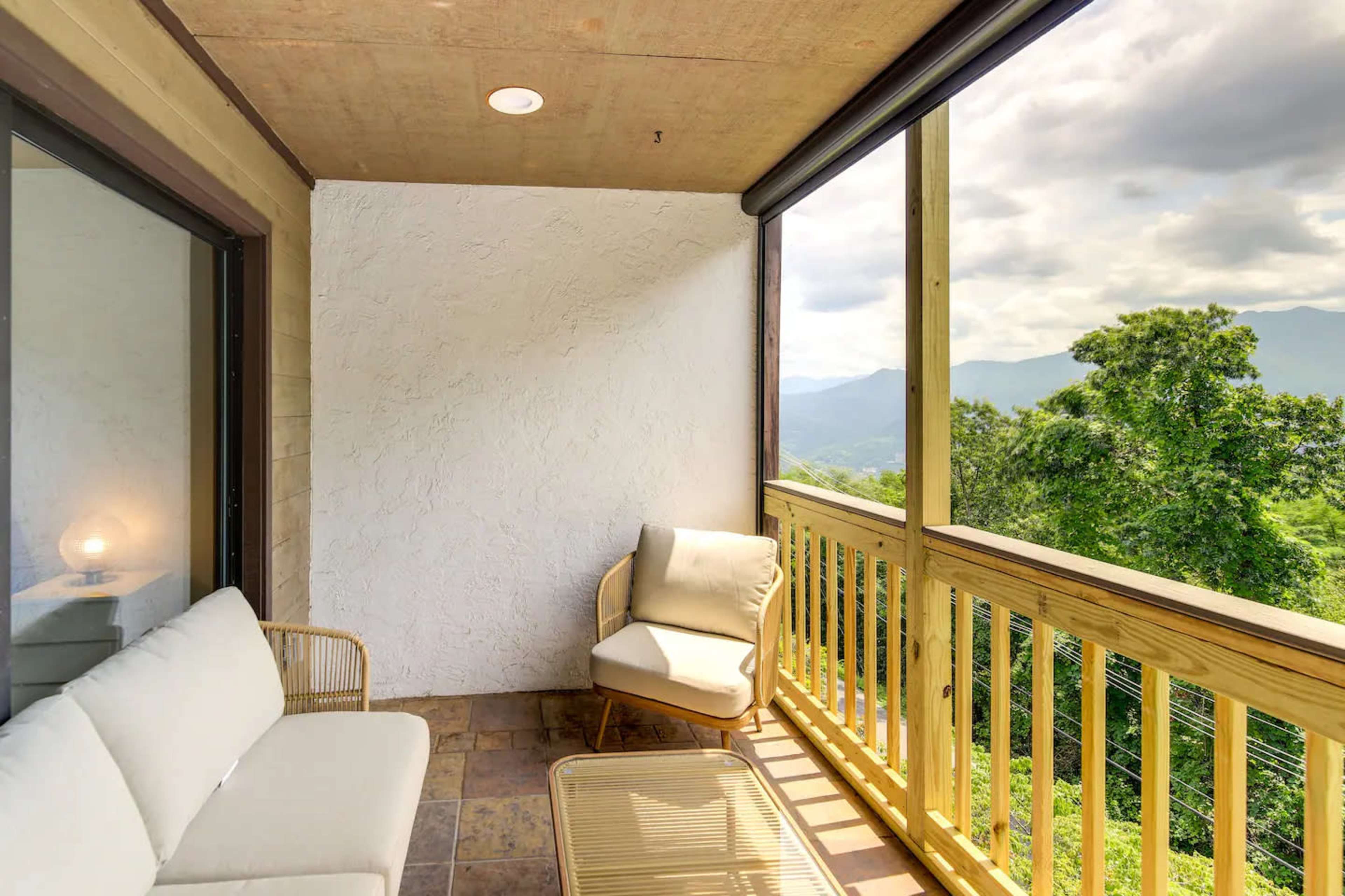 The image shows a balcony with a seating area, featuring a light-colored sofa, a single chair, and a view of distant mountains and trees.