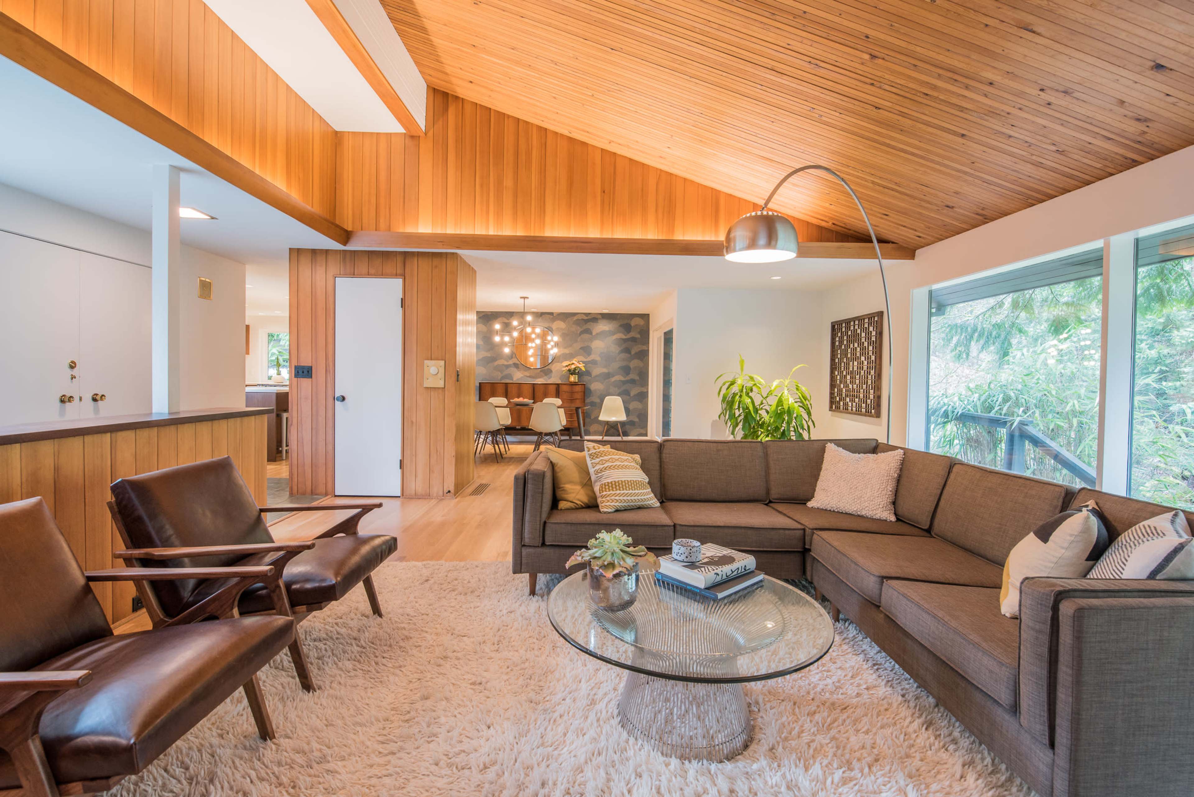 A modern living room with a wooden ceiling, a sectional sofa, a glass coffee table, and two leather armchairs, all situated near large windows.