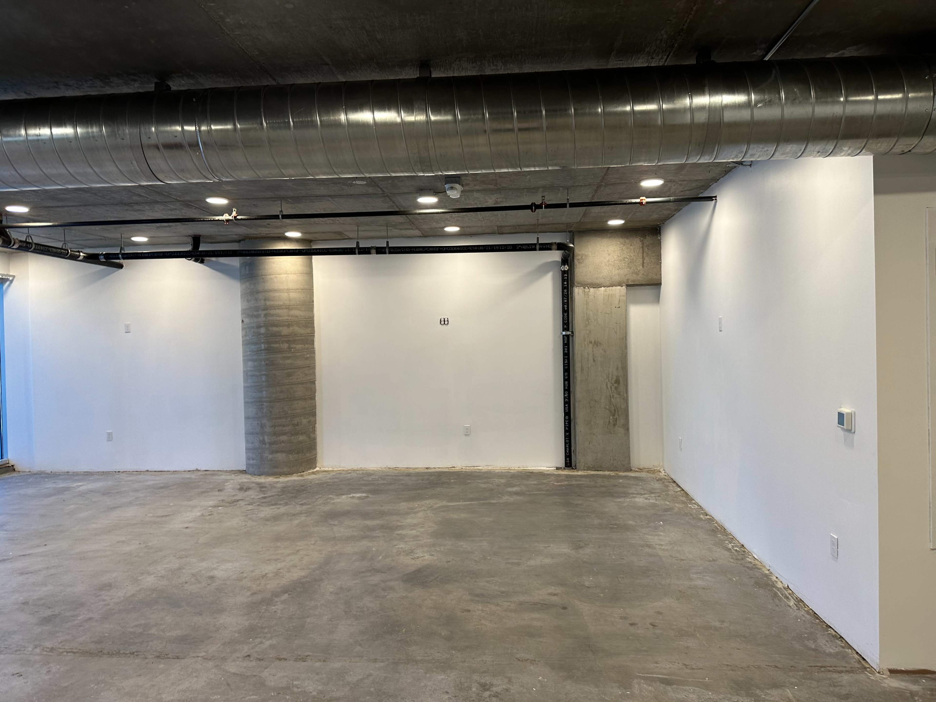 The image shows an unfinished interior space with bare concrete floors and white walls, featuring exposed ductwork and a doorway at the far end.