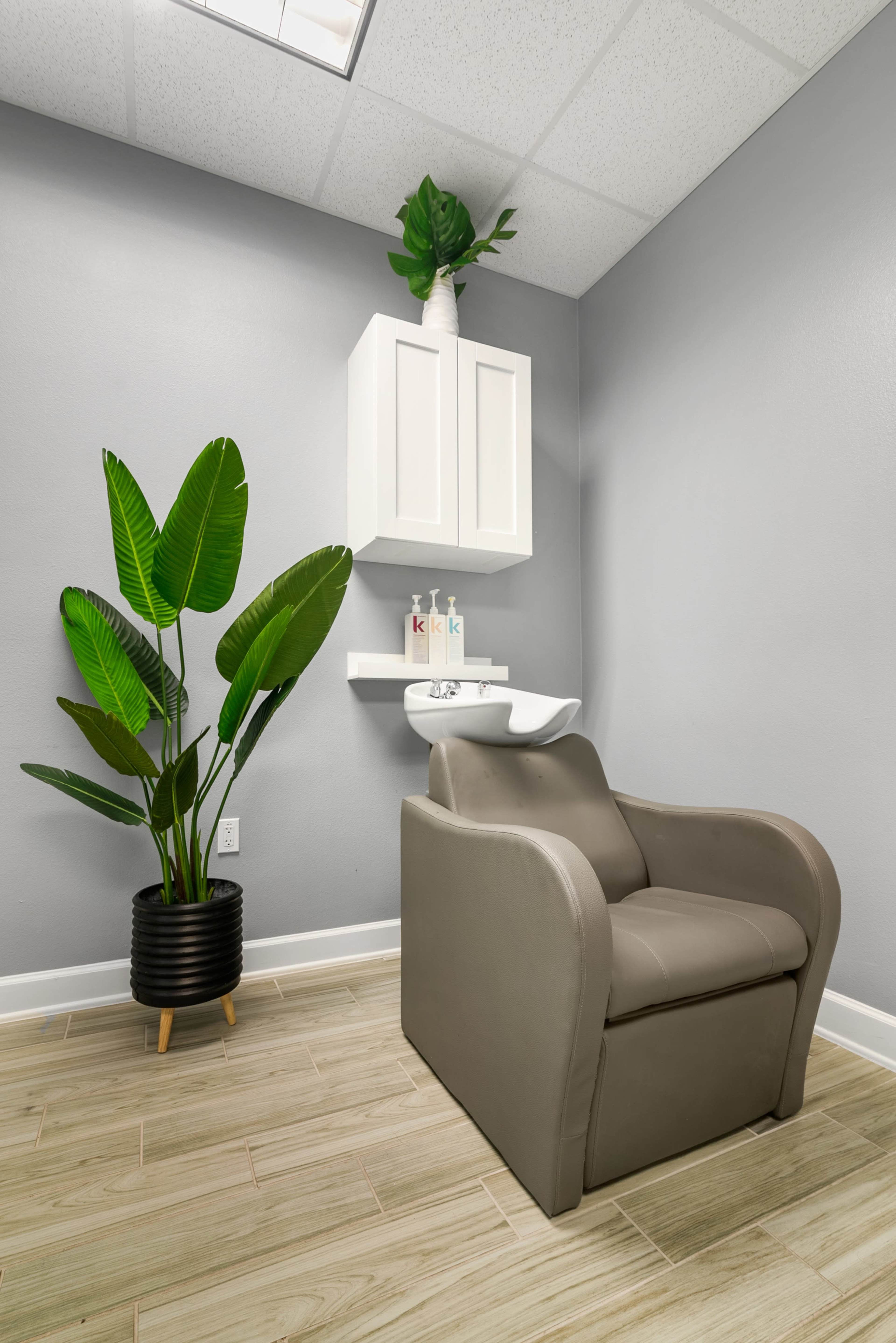 A small salon corner featuring a gray chair, a white cabinet mounted on the wall, and a potted plant in a black pot.