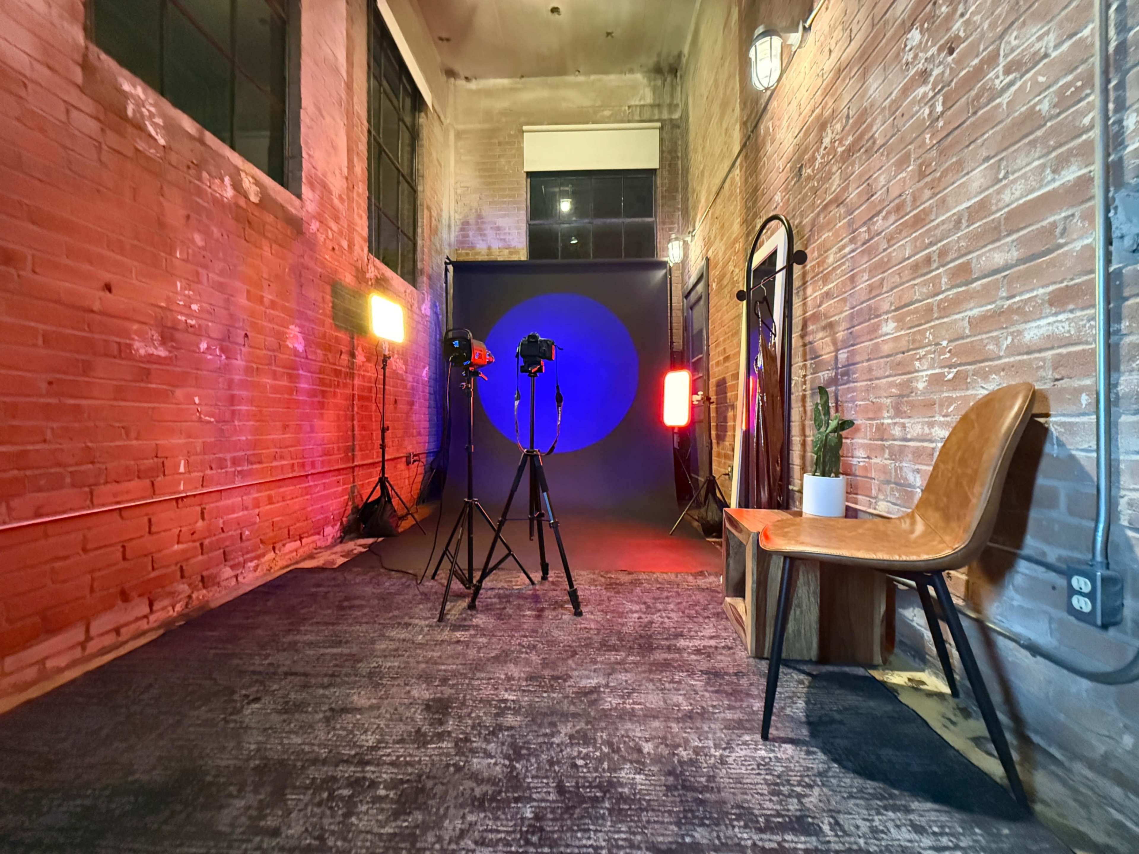 The image shows a photography studio setup with two lights, a backdrop, and a chair beside a mirror along a brick wall.