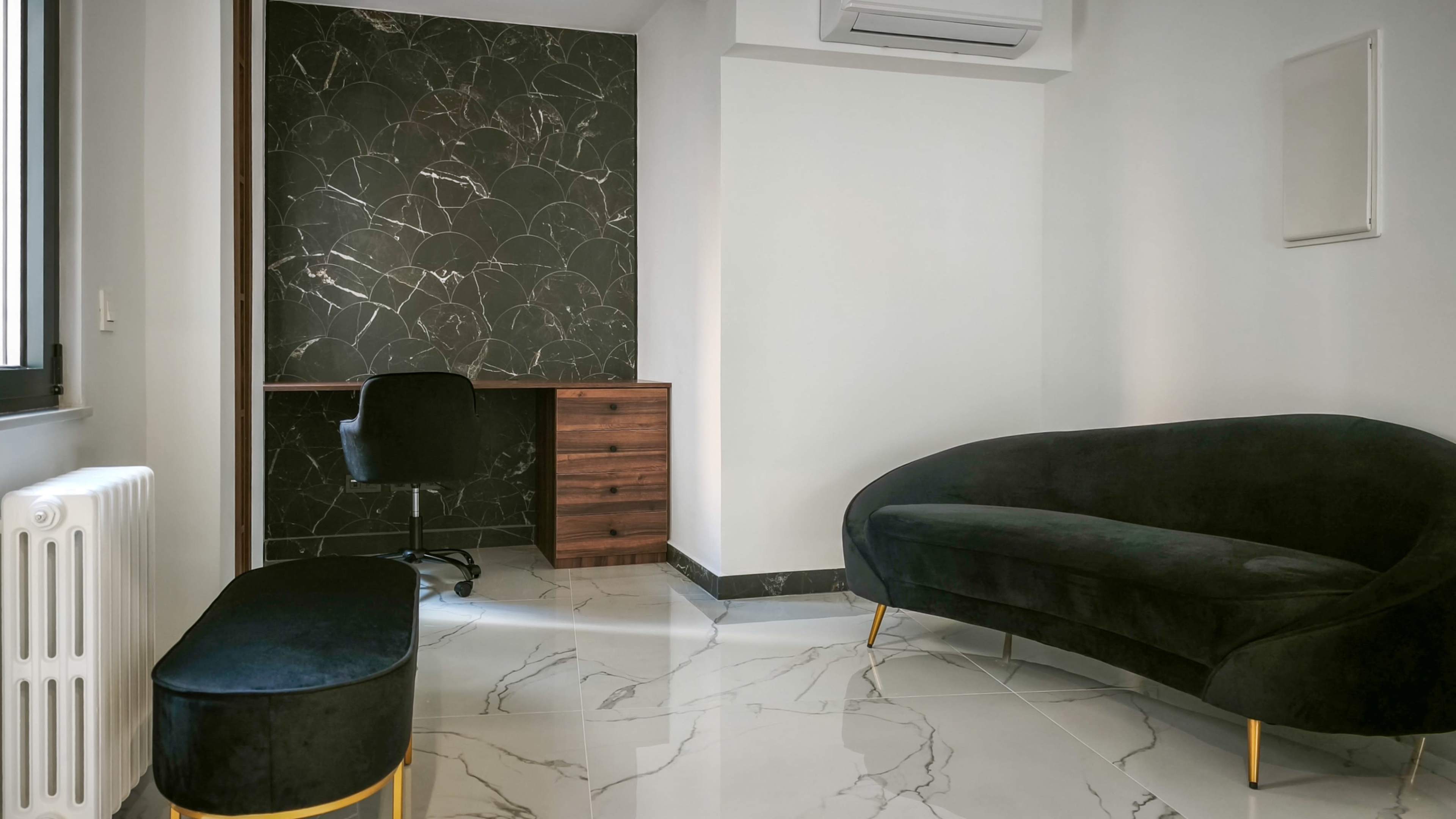 The image shows a modern room with a black marble accent wall, a wooden desk with a chair, a dark green sofa, and a matching ottoman on a marble floor.