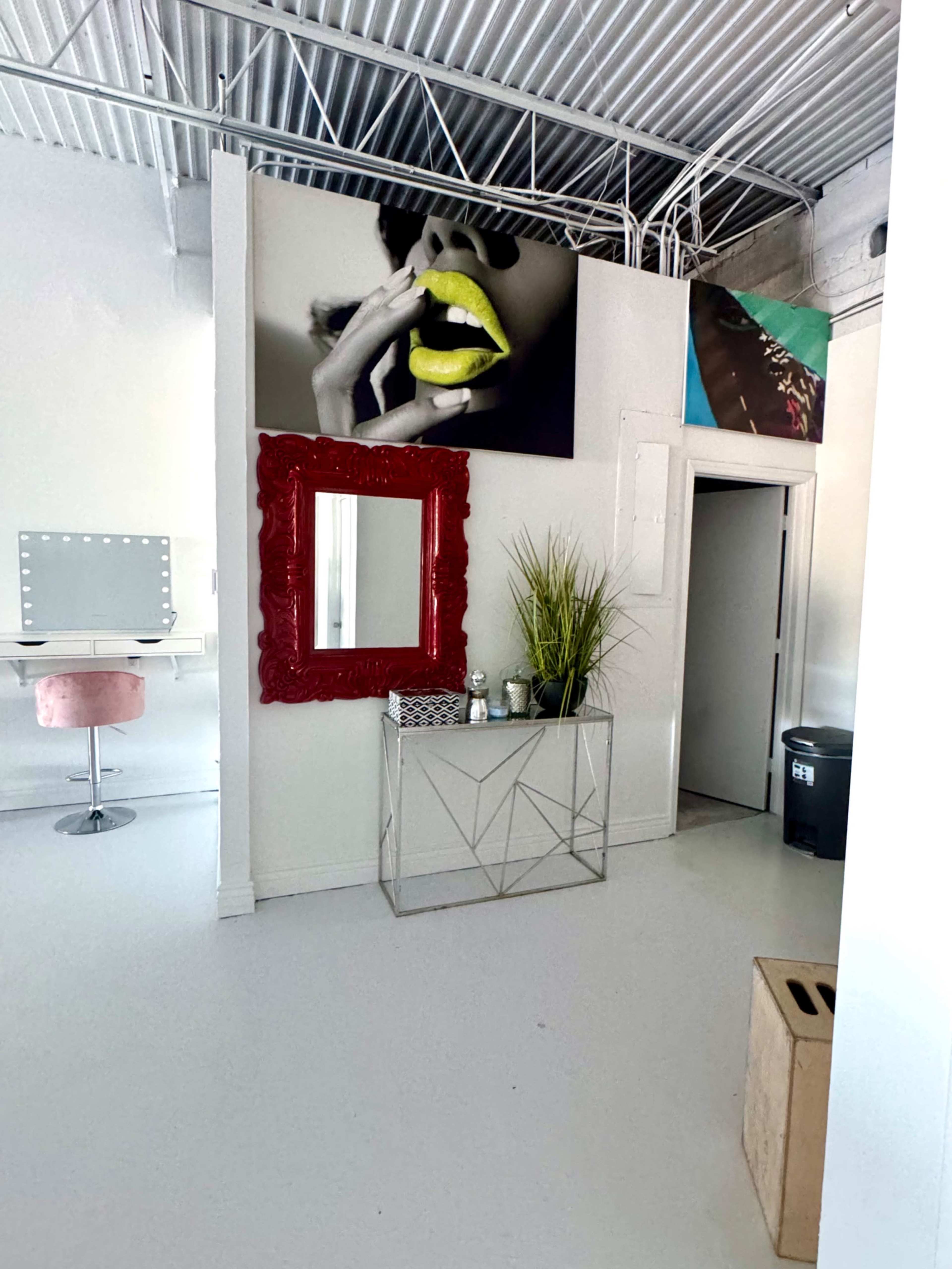 The image shows a modern, minimalist interior featuring a decorative red mirror, a silver geometric console table, and a large photograph on the wall, alongside a doorway and a small vanity area with a plush pink stool.