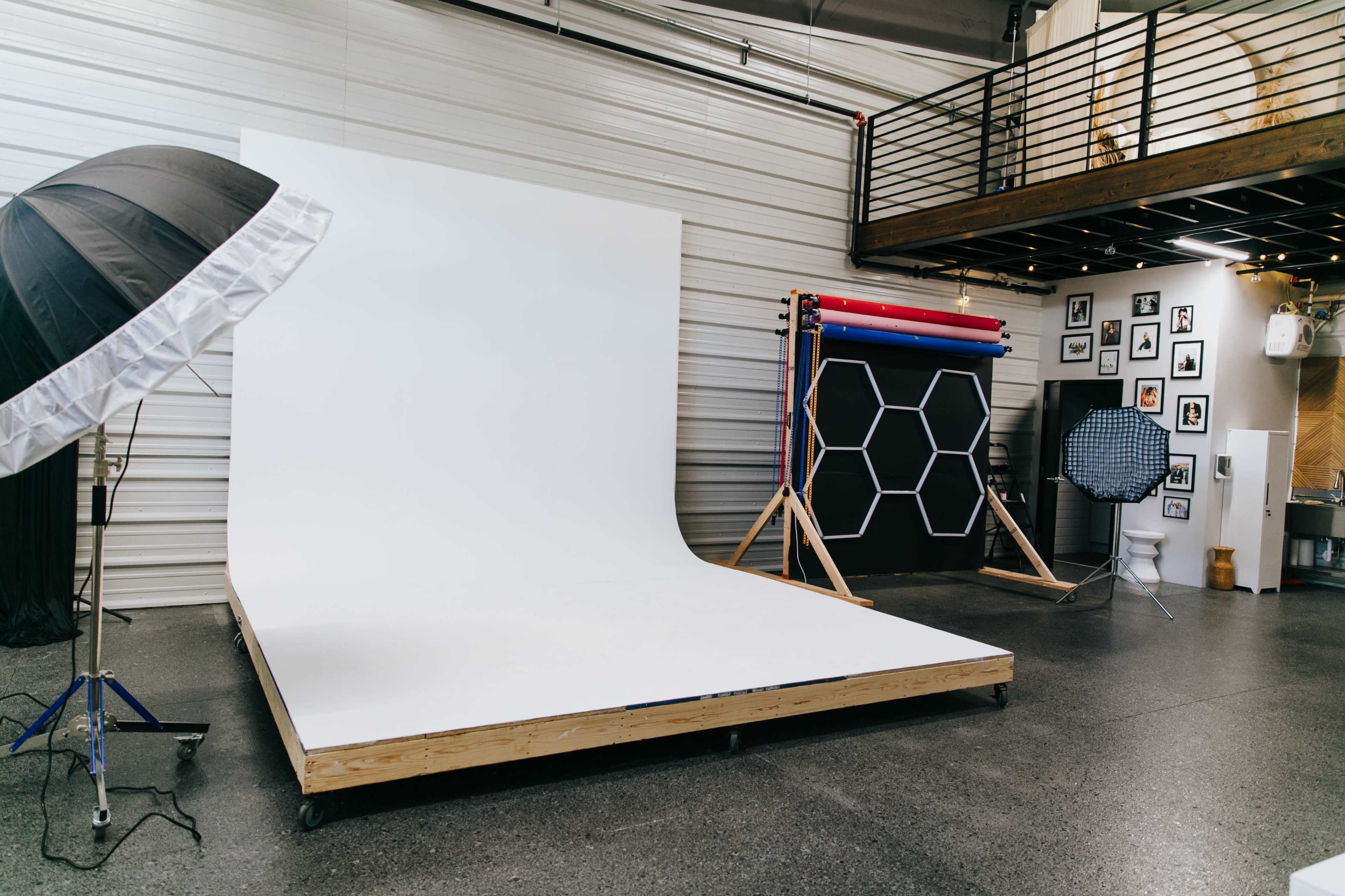 The image shows a photography studio with a white backdrop, various lighting equipment, and a display of colorful backdrops mounted on a stand.