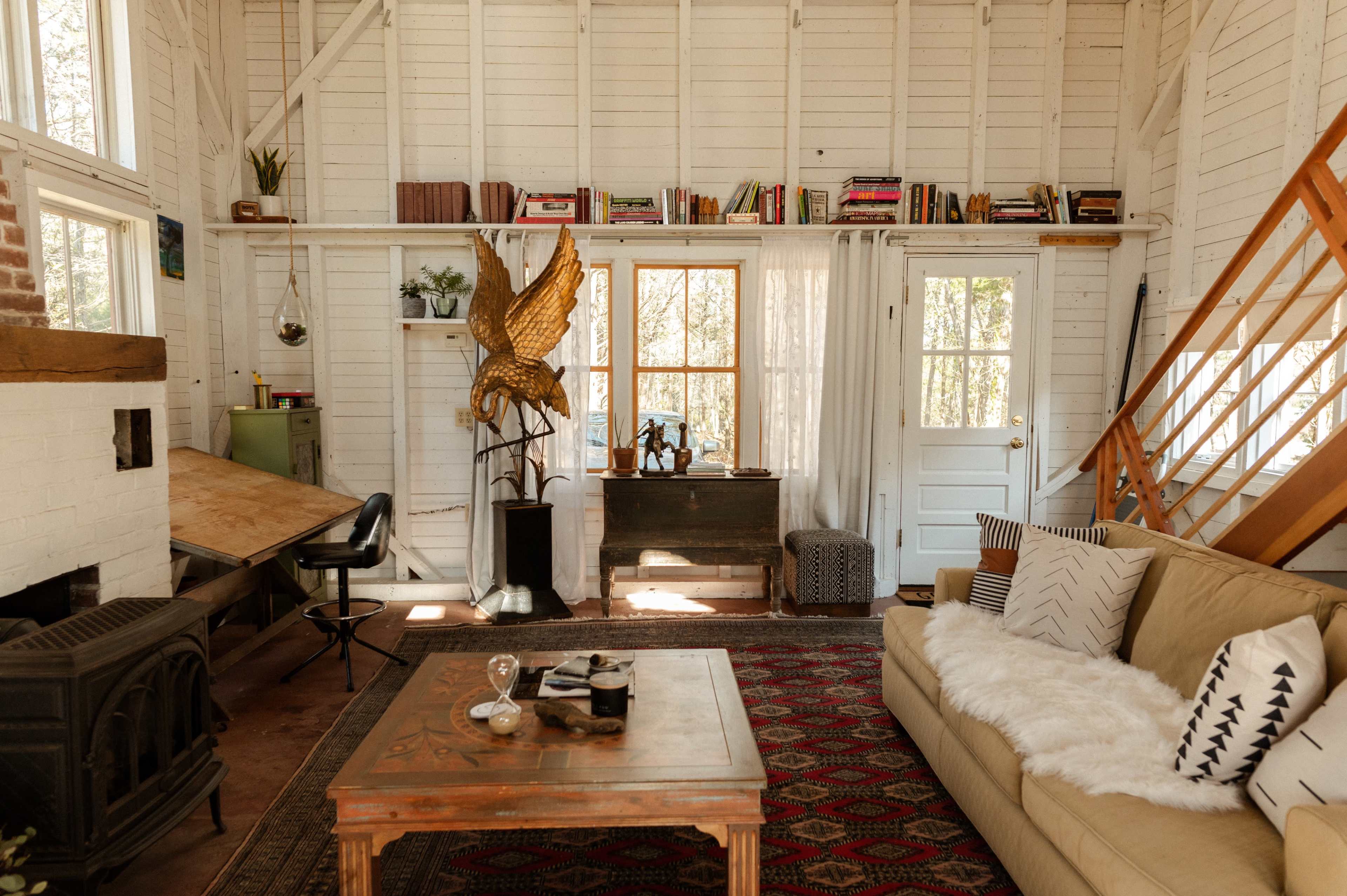 The image shows a cozy living space with a beige sofa, a wooden table for work, and bookshelves lined with books, accented by a golden sculpture and a large window allowing natural light.