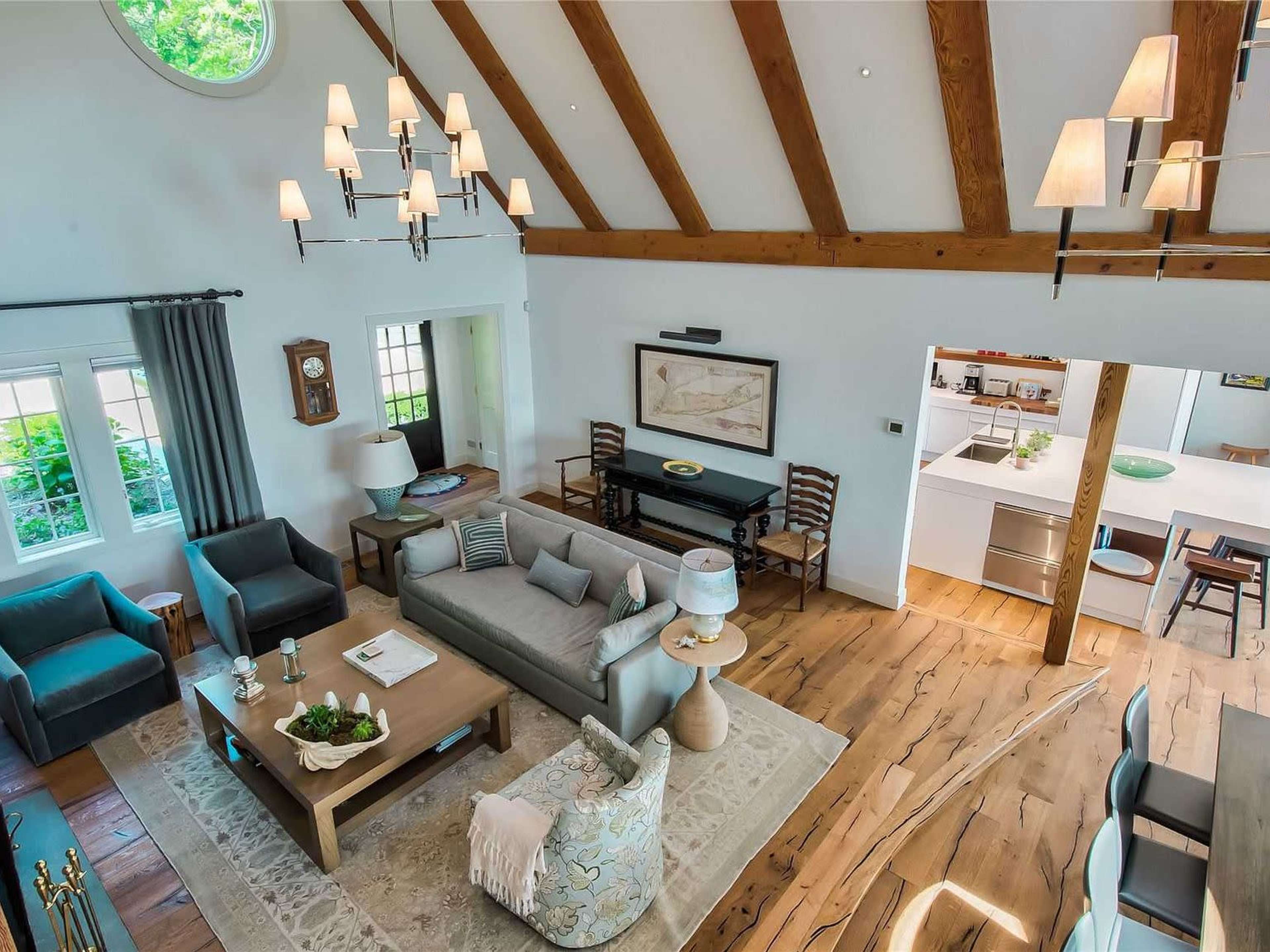 The image shows a spacious living room with vaulted ceilings, wooden beams, and modern furniture, including a gray sofa and several seating areas.