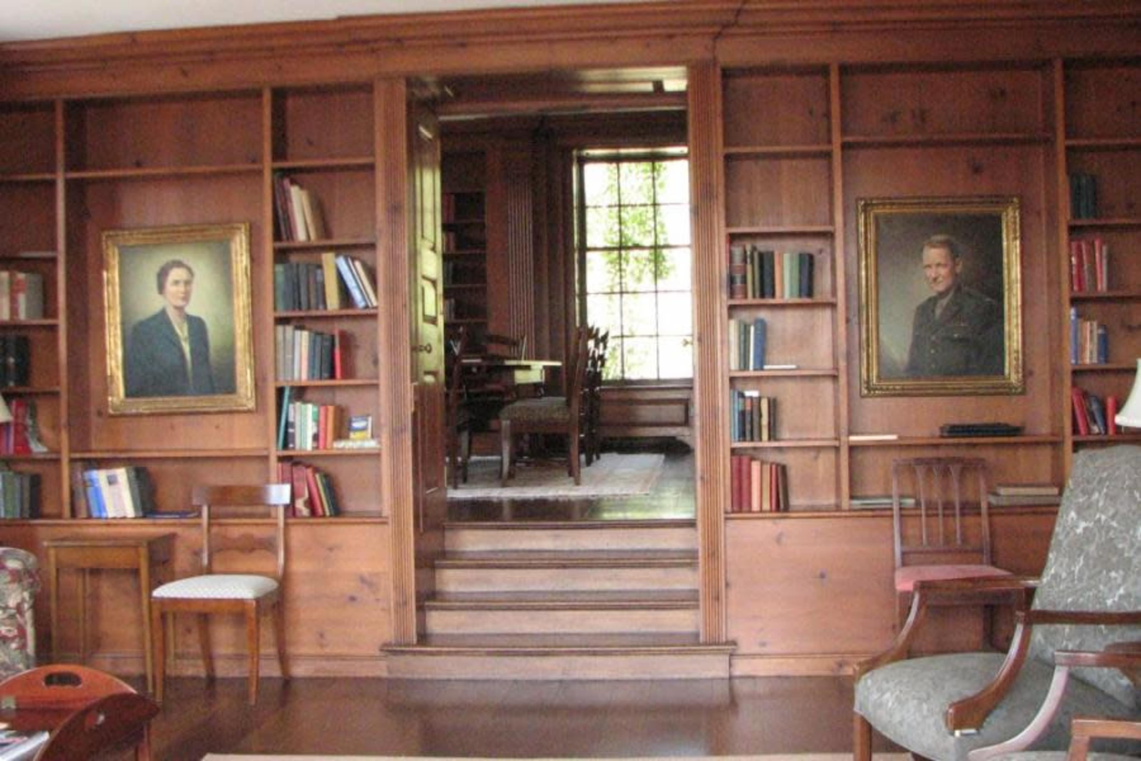 The image shows a wooden library with bookshelves lining the walls, two framed portraits hanging, and a staircase leading to a room with a dining area visible in the background.