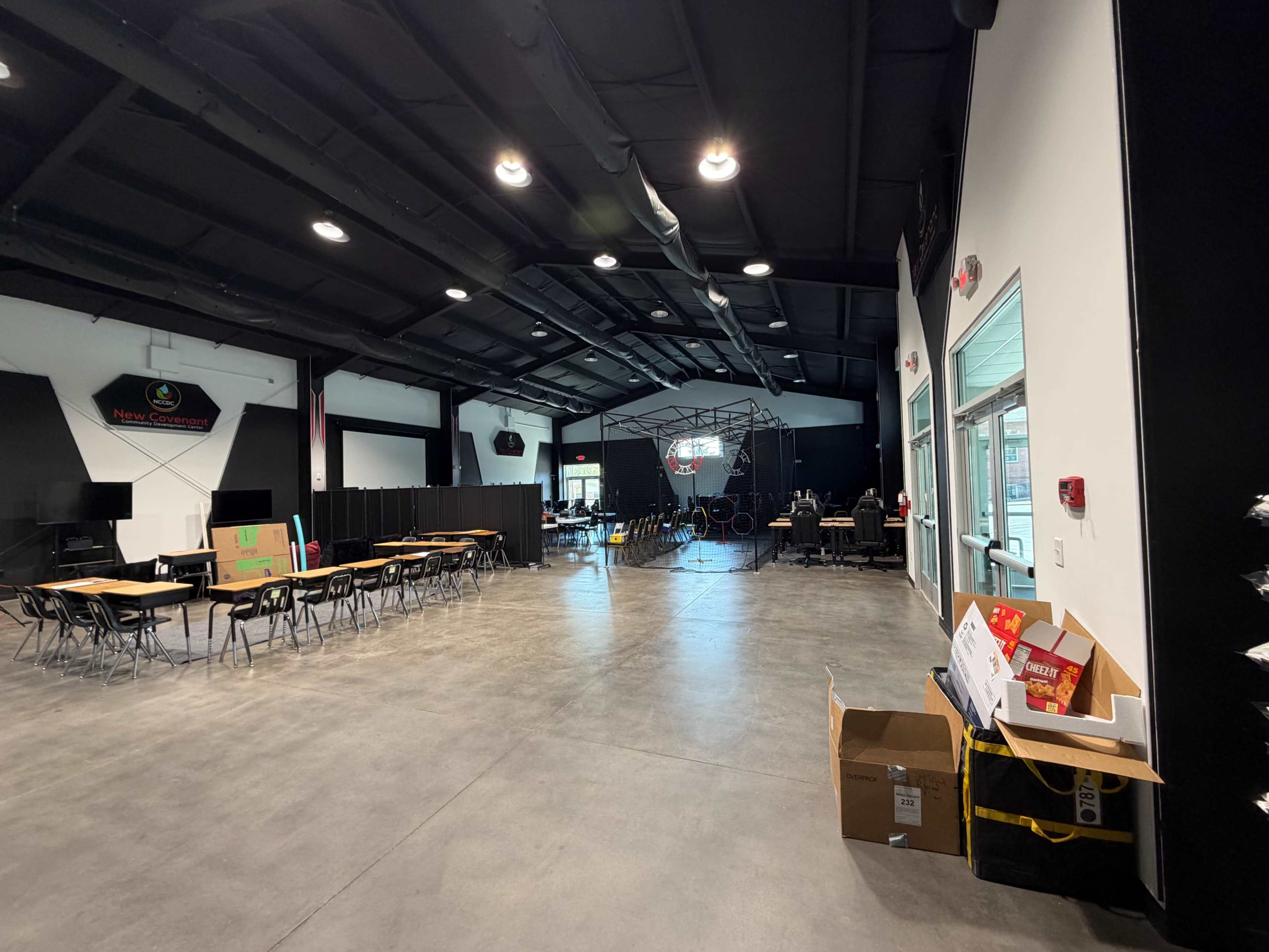 The image shows a spacious indoor area with tables and chairs arranged on one side, and a stage-like setup with black curtains on the opposite side.