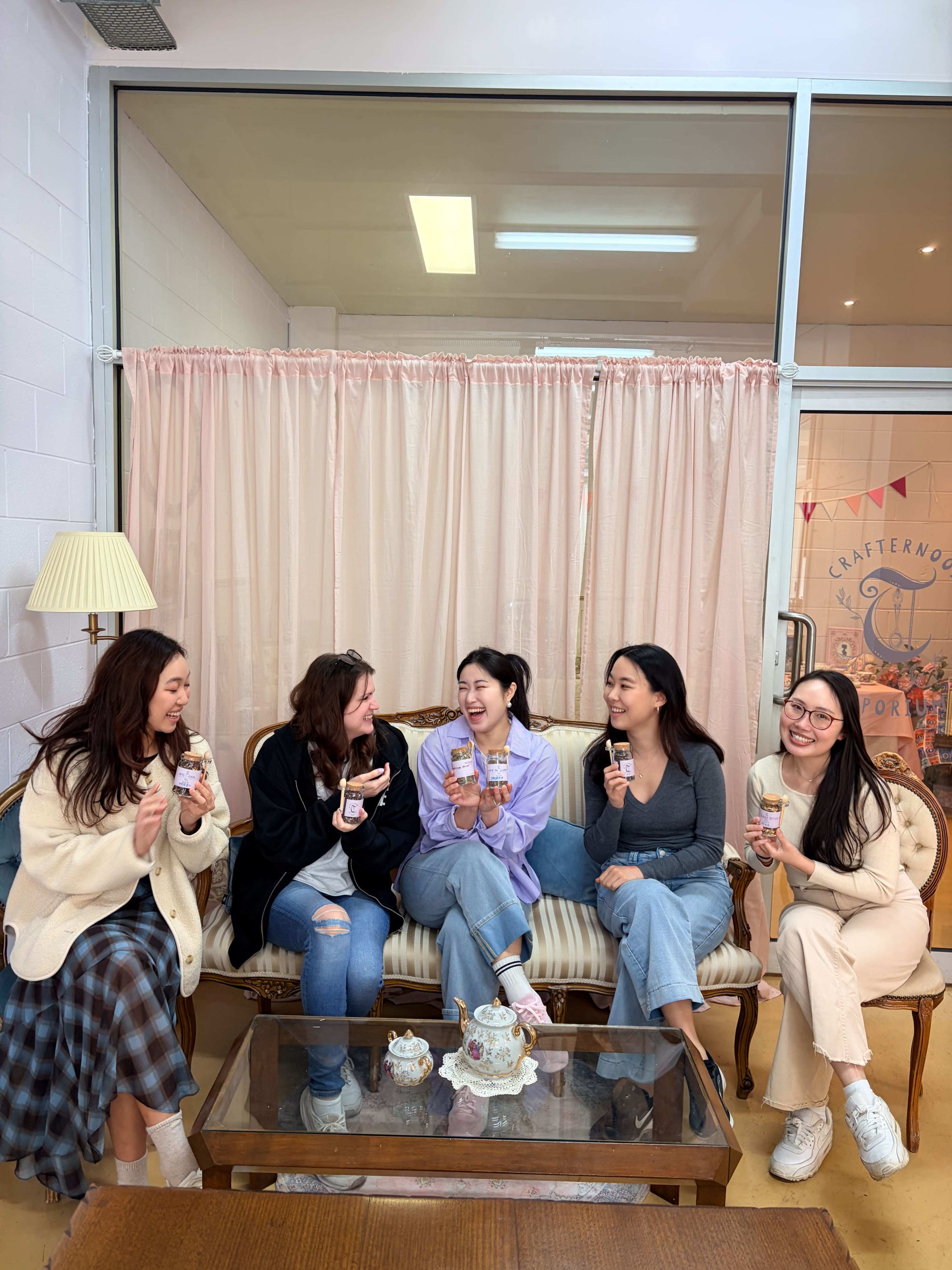 A group of five young women sit together on a couch, holding dessert cups and smiling, in a cozy room decorated with soft pastel colors.