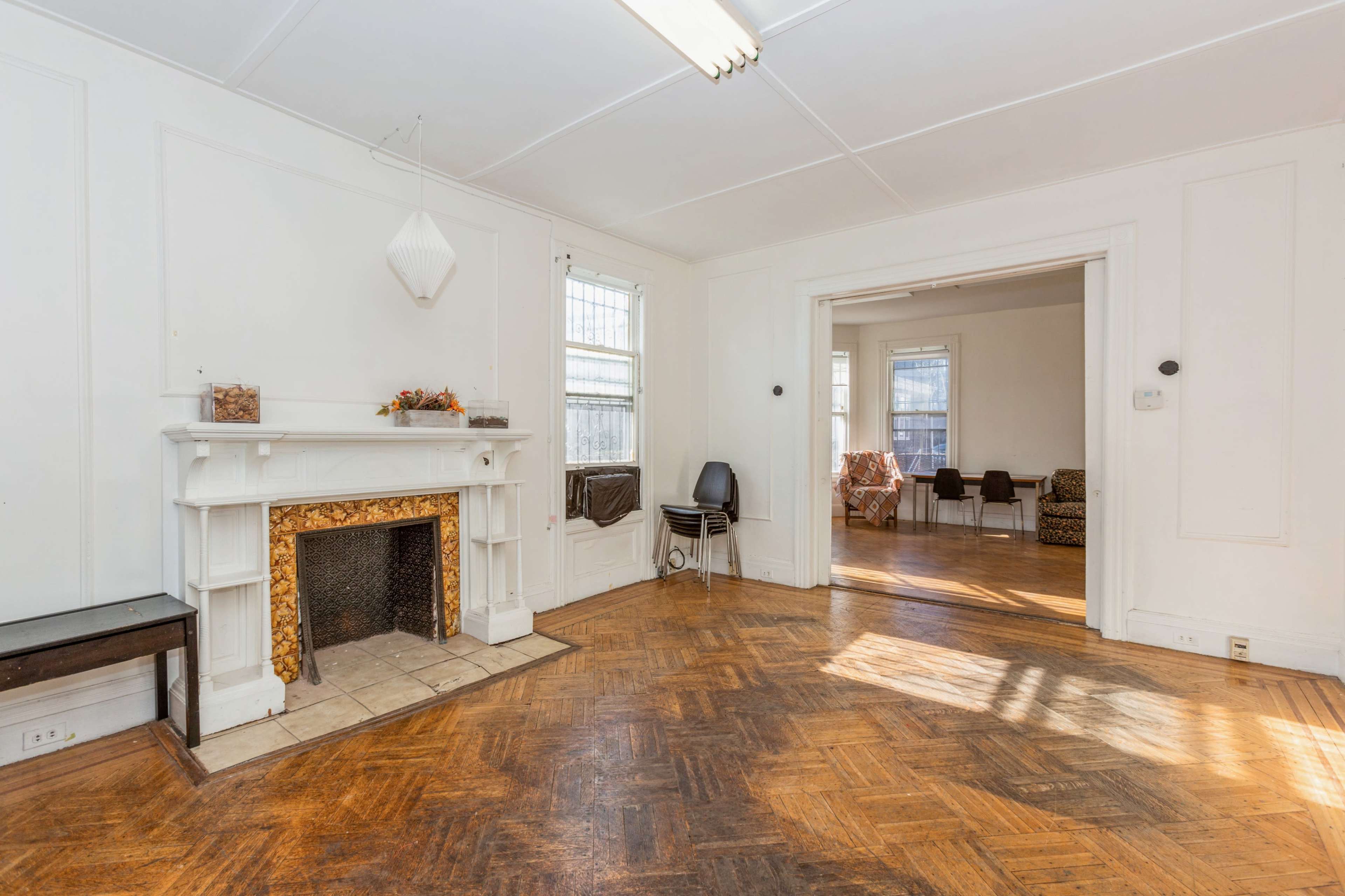 The image shows a spacious room with a decorative fireplace and hardwood flooring, leading to an adjacent room furnished with chairs.