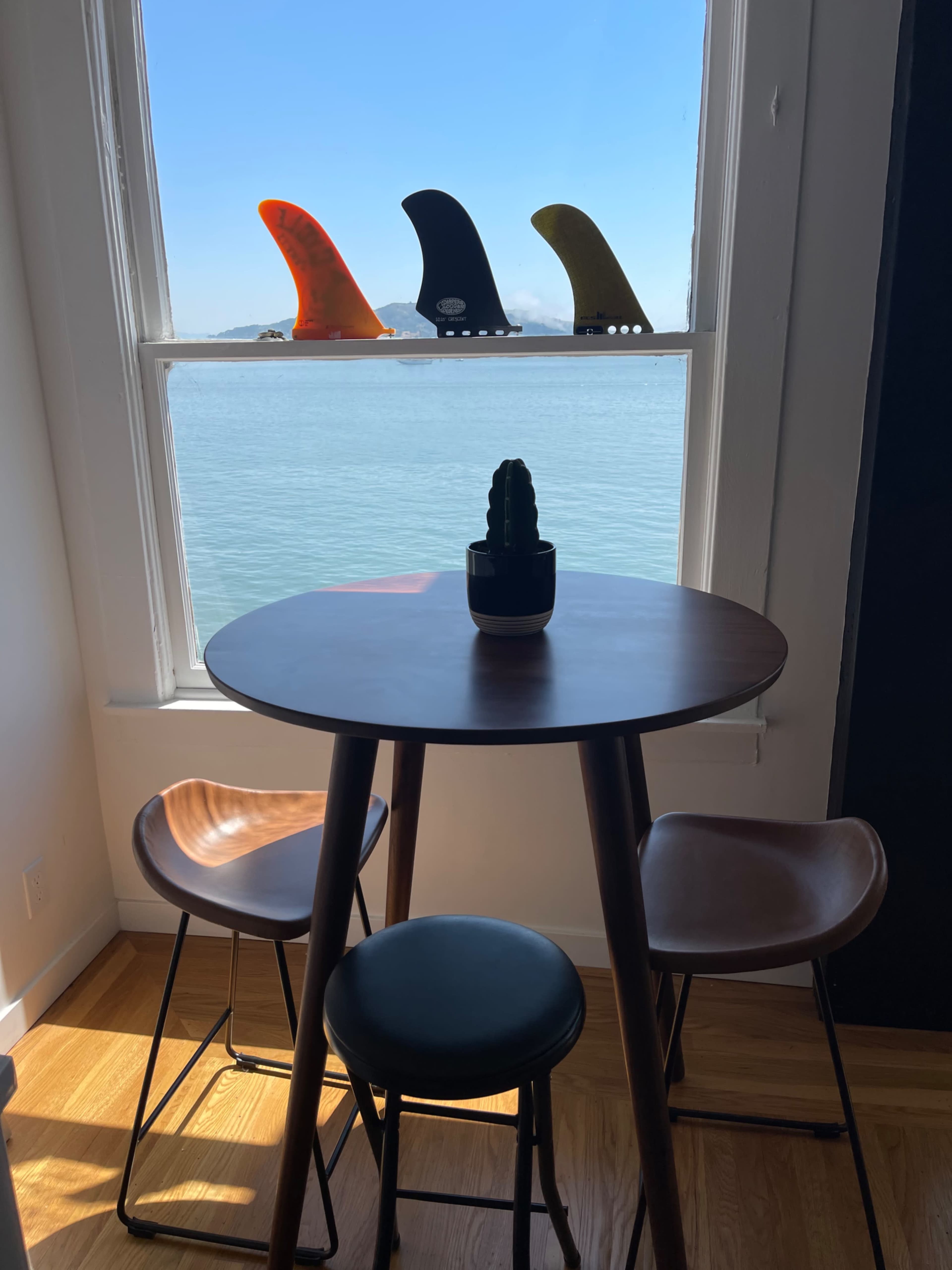 A small table with two chairs is positioned by a window overlooking a body of water, with colorful surfboard fins displayed on the windowsill.