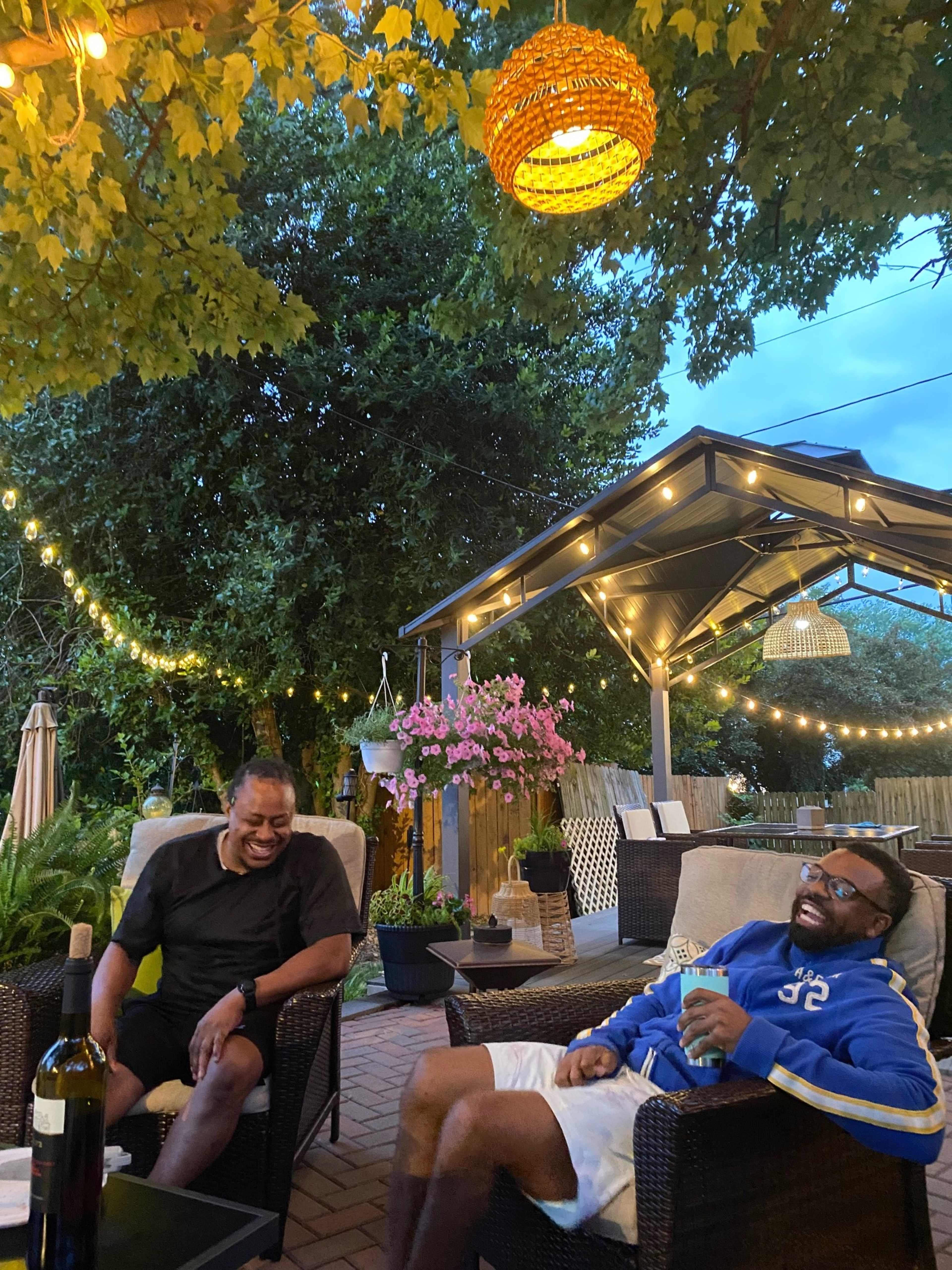 Two men are sitting in a garden area with string lights overhead, surrounded by plants and flowers, and a bottle of wine is on the table between them.