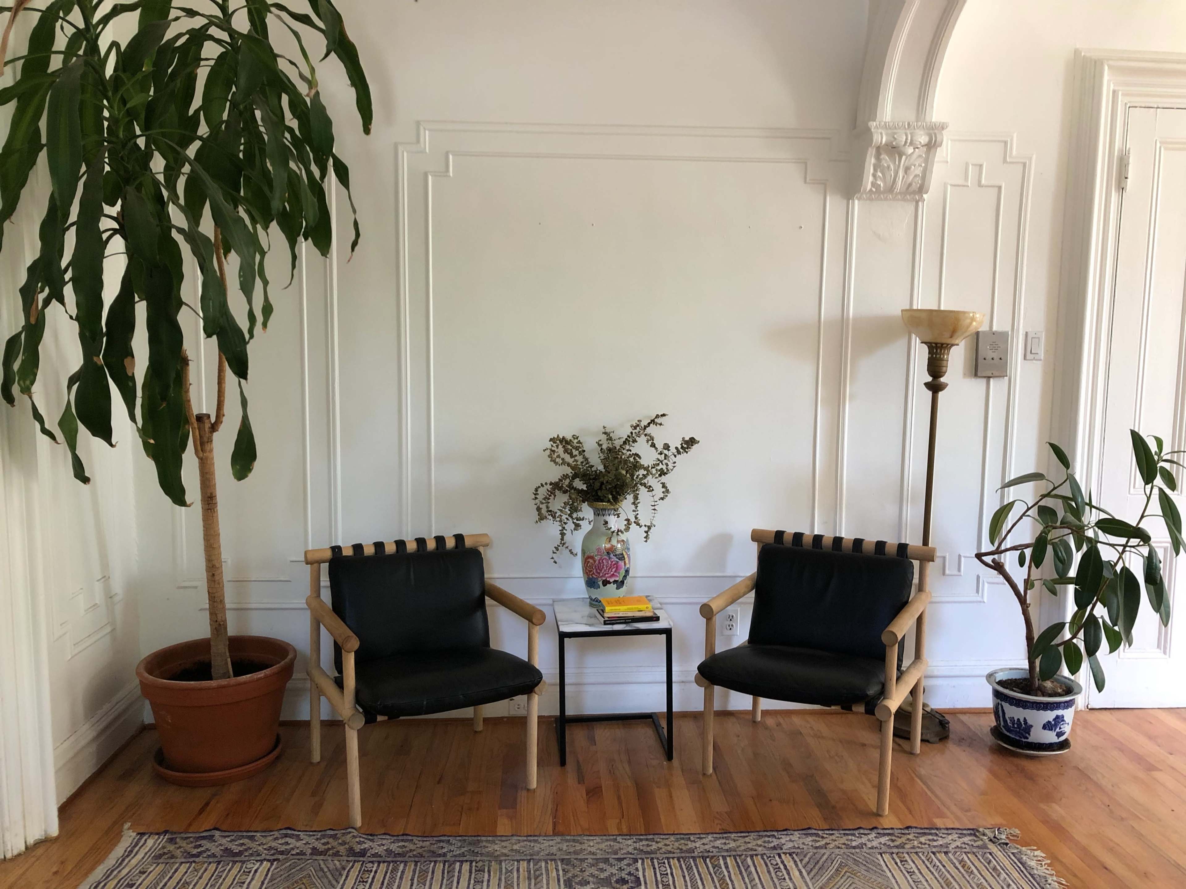 A minimalist interior space with two black chairs, a small side table, a vase of plants, and two potted trees flanking the seating area.