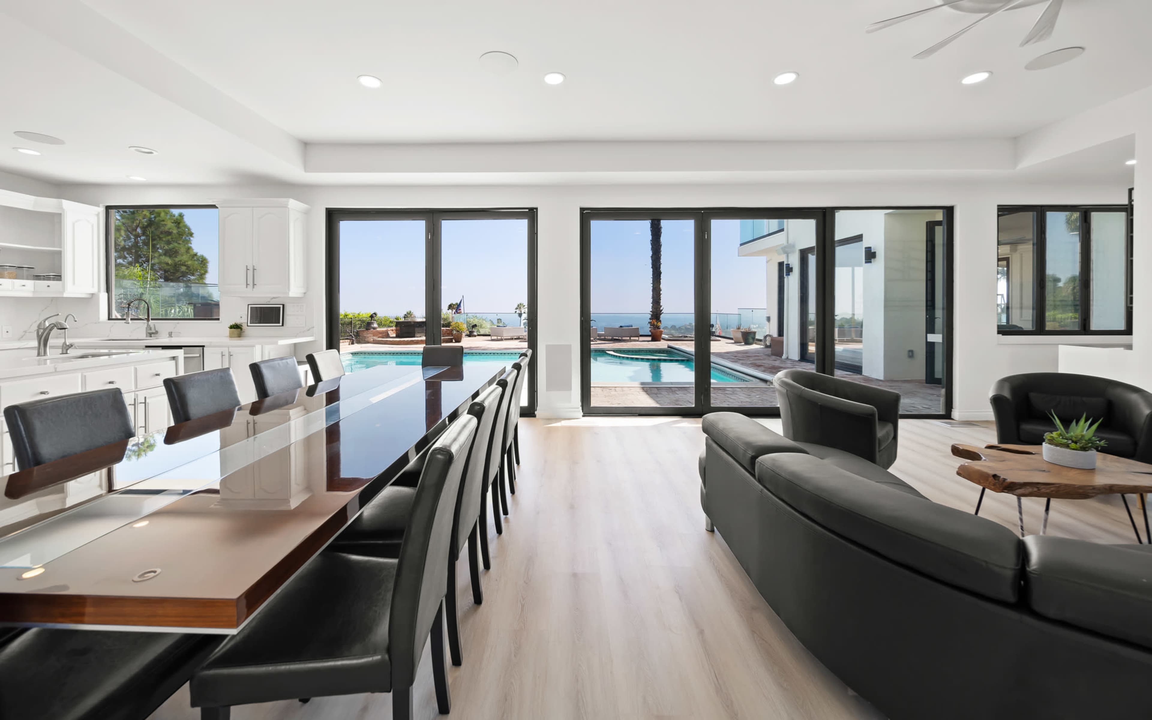 A modern open-concept living space features a large dining table, black leather seating, and large glass doors leading to a pool area with views of the ocean.