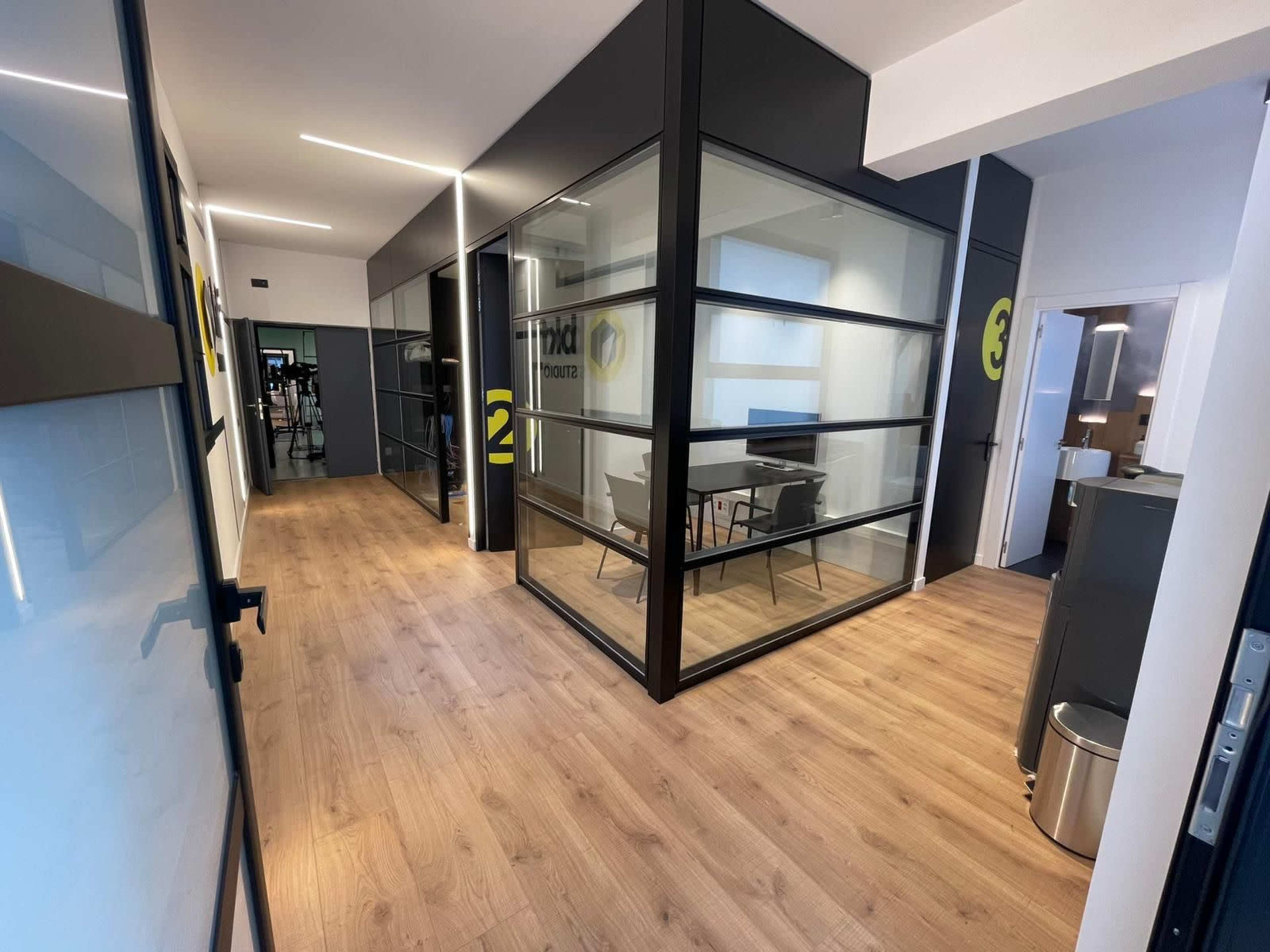 A modern office corridor with glass-enclosed meeting rooms on one side and a clean, wooden floor.
