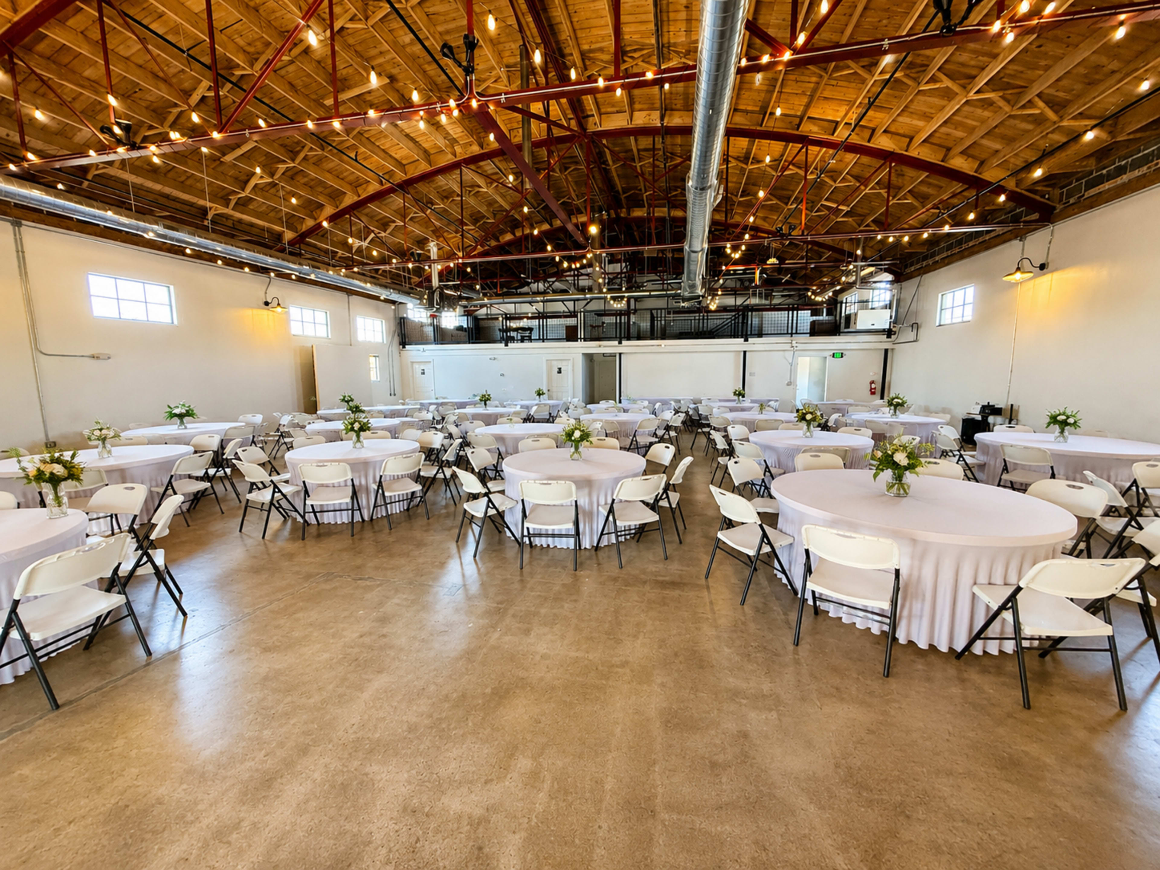 The image shows a spacious event venue with circular tables covered in light-colored tablecloths, each adorned with small flower arrangements, set up for a gathering.