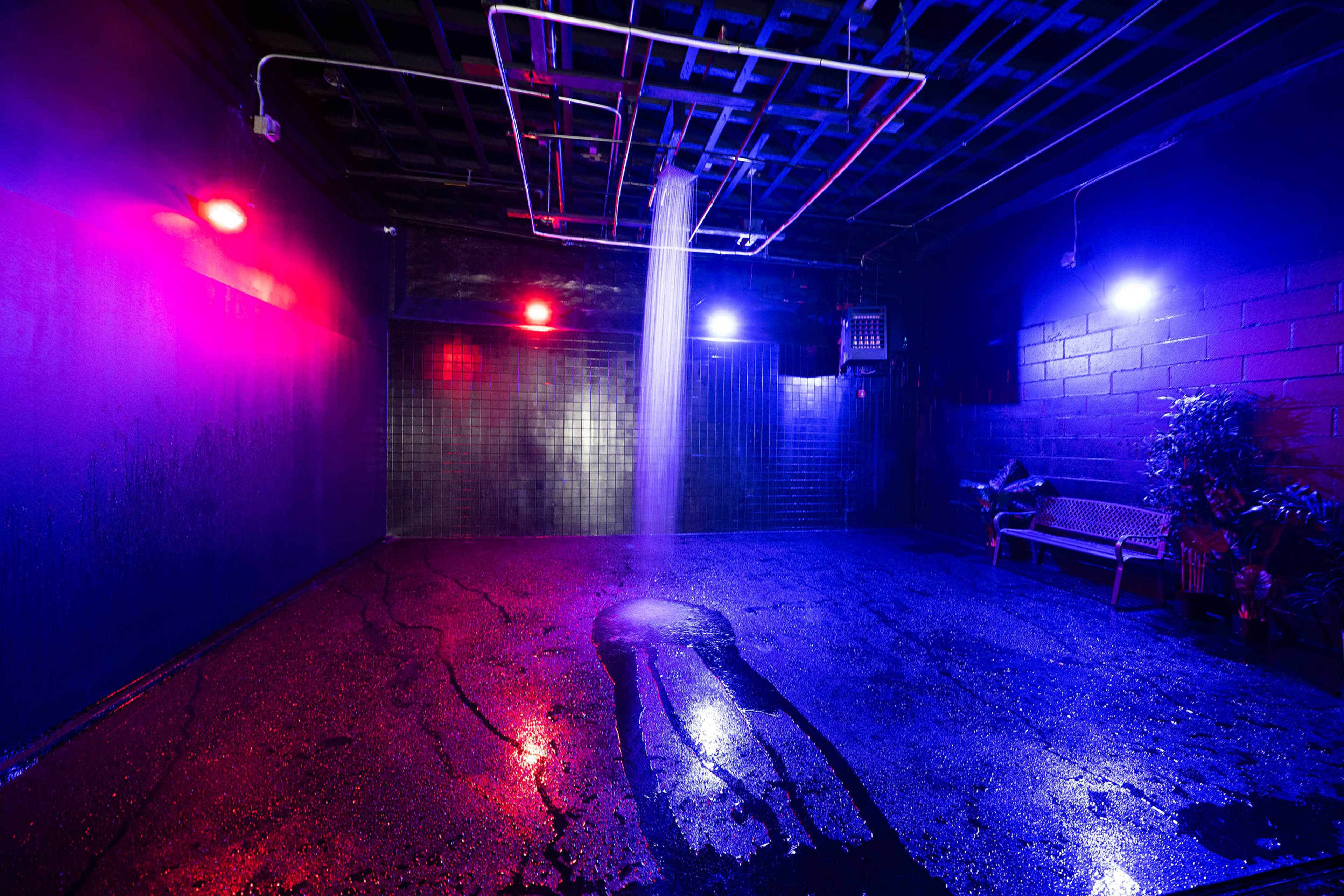 A dimly lit room features hanging pipes from which water flows onto a shiny, wet floor, illuminated by purple and blue lights.