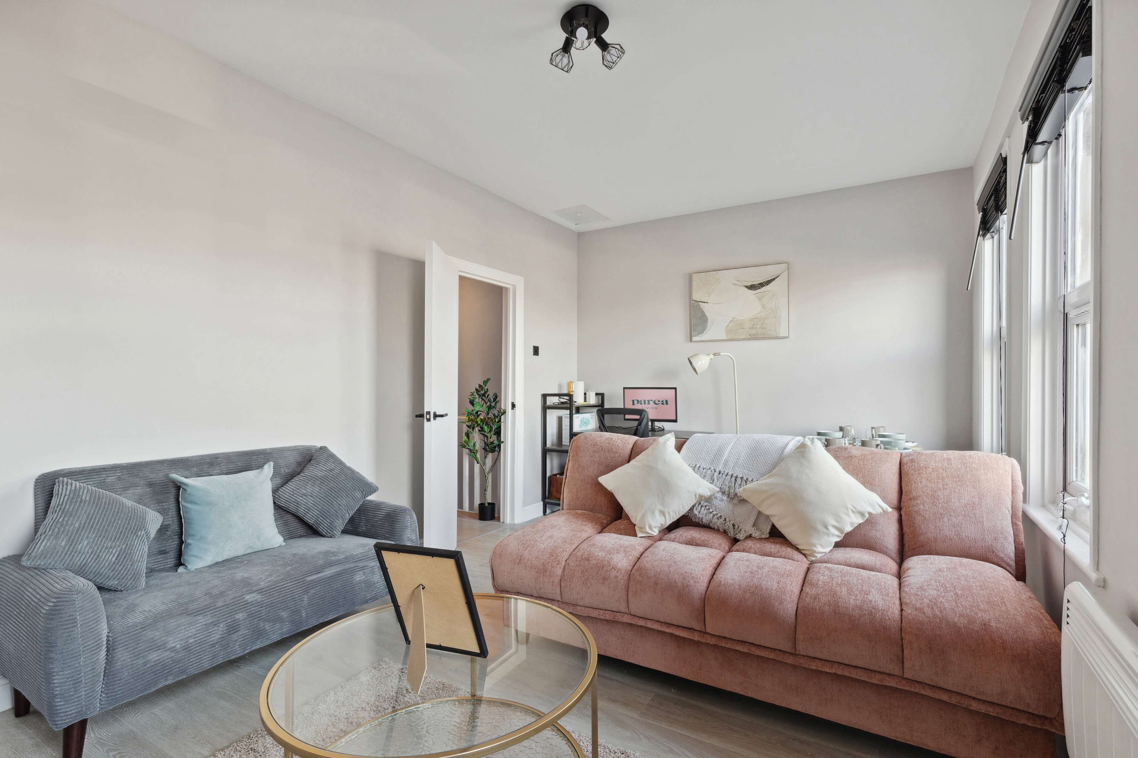 A well-lit living room features a gray sofa and a pink couch with decorative pillows, accompanied by a glass coffee table and a plant in the corner.