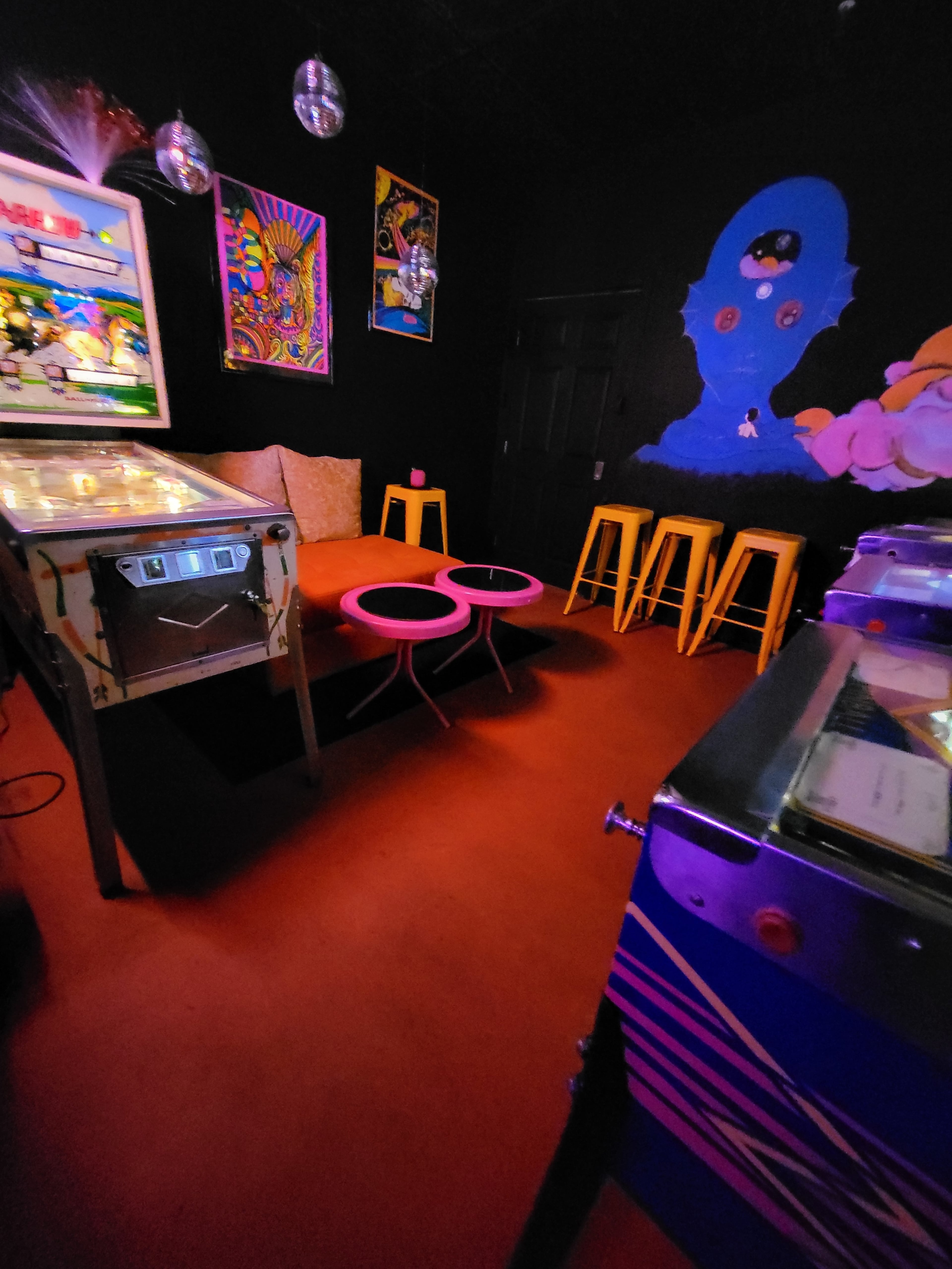 The scene features a small arcade room with pinball machines, vibrant wall art, a pair of pink tables, and yellow stools against a black backdrop.