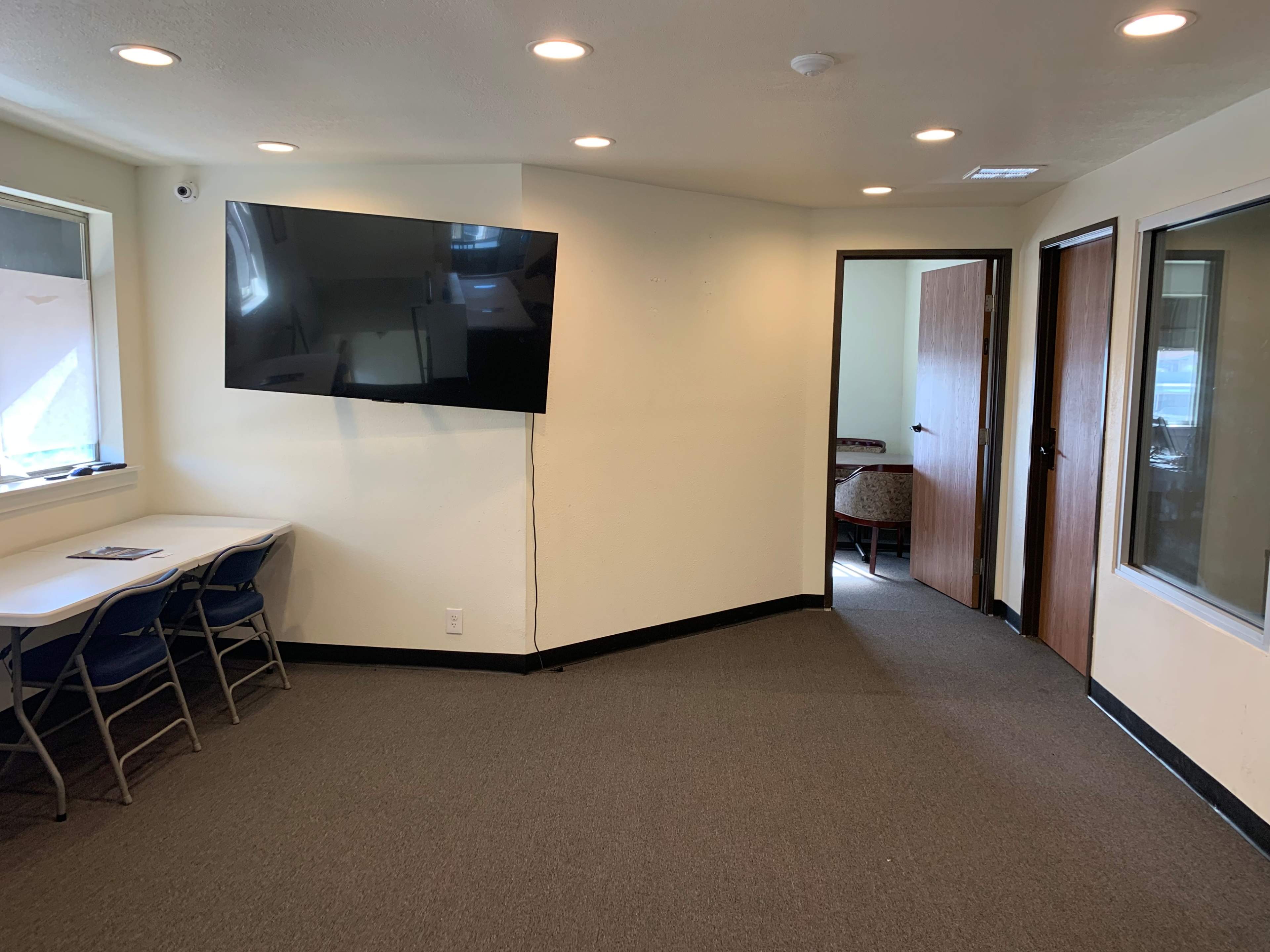 A small meeting room with a large wall-mounted TV, a table with chairs, and a door leading to another room.