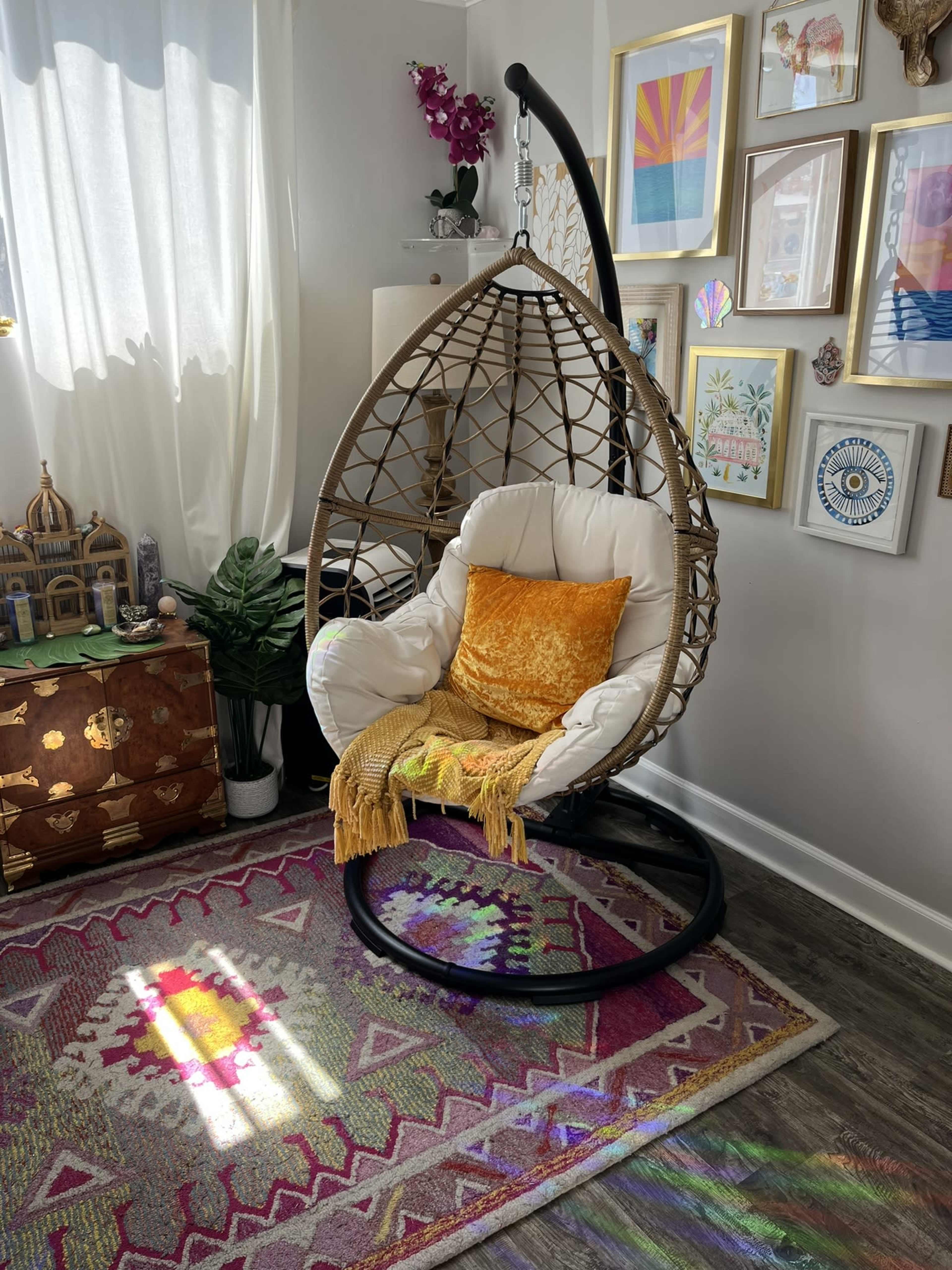 A wicker hanging chair with a white cushion and orange throw pillow is positioned in a brightly lit room adorned with wall art and a colorful rug.