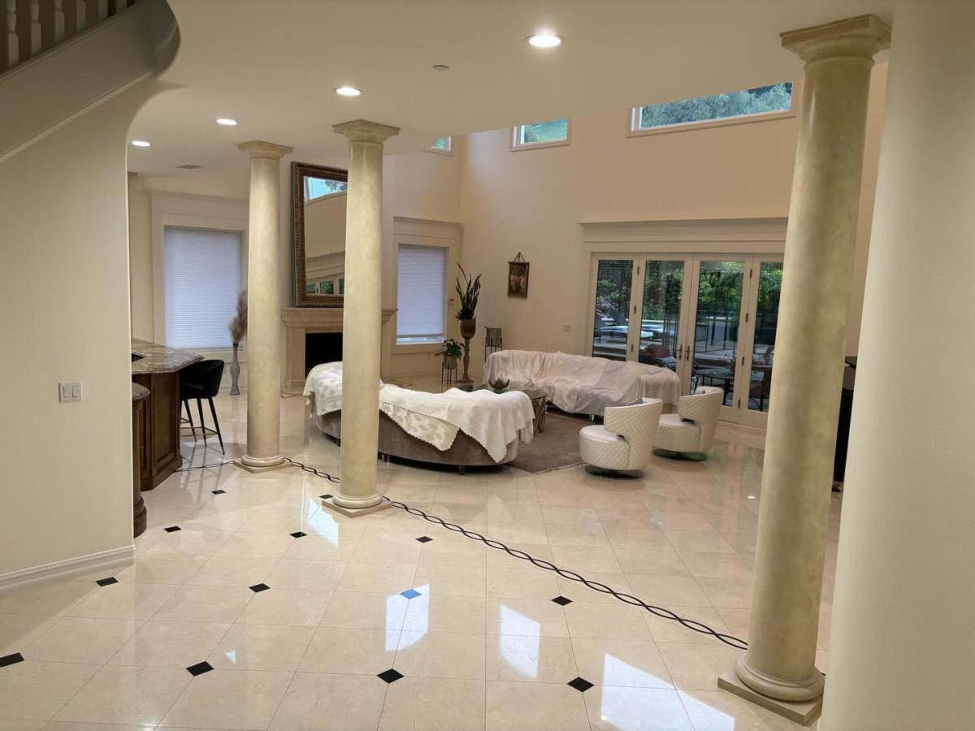 The image shows a spacious interior living area featuring marble flooring, large windows, and white furniture arranged around a fireplace.