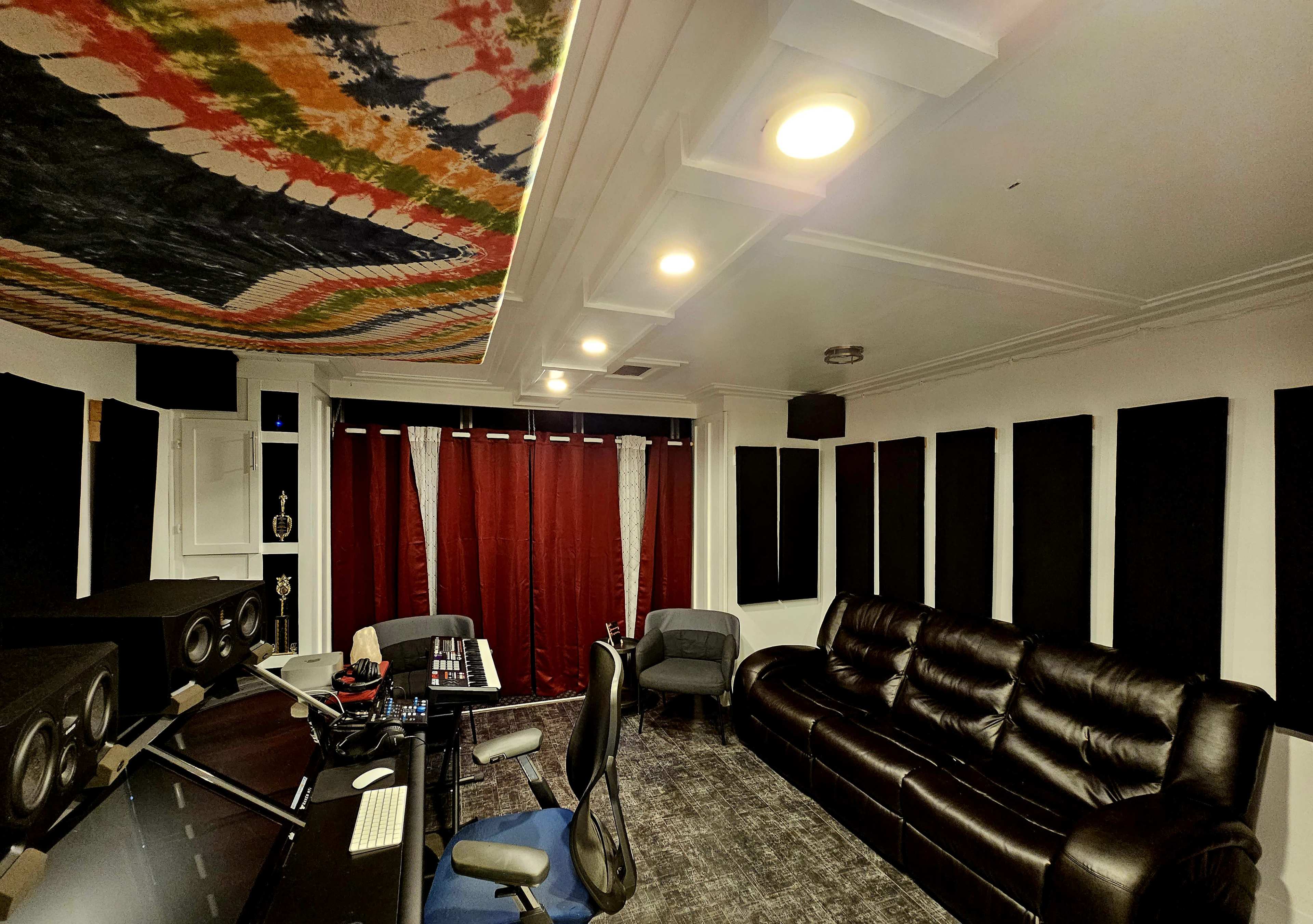 A sound studio with a black leather sofa, acoustic panels on the walls, a control desk with audio equipment, and red curtains.