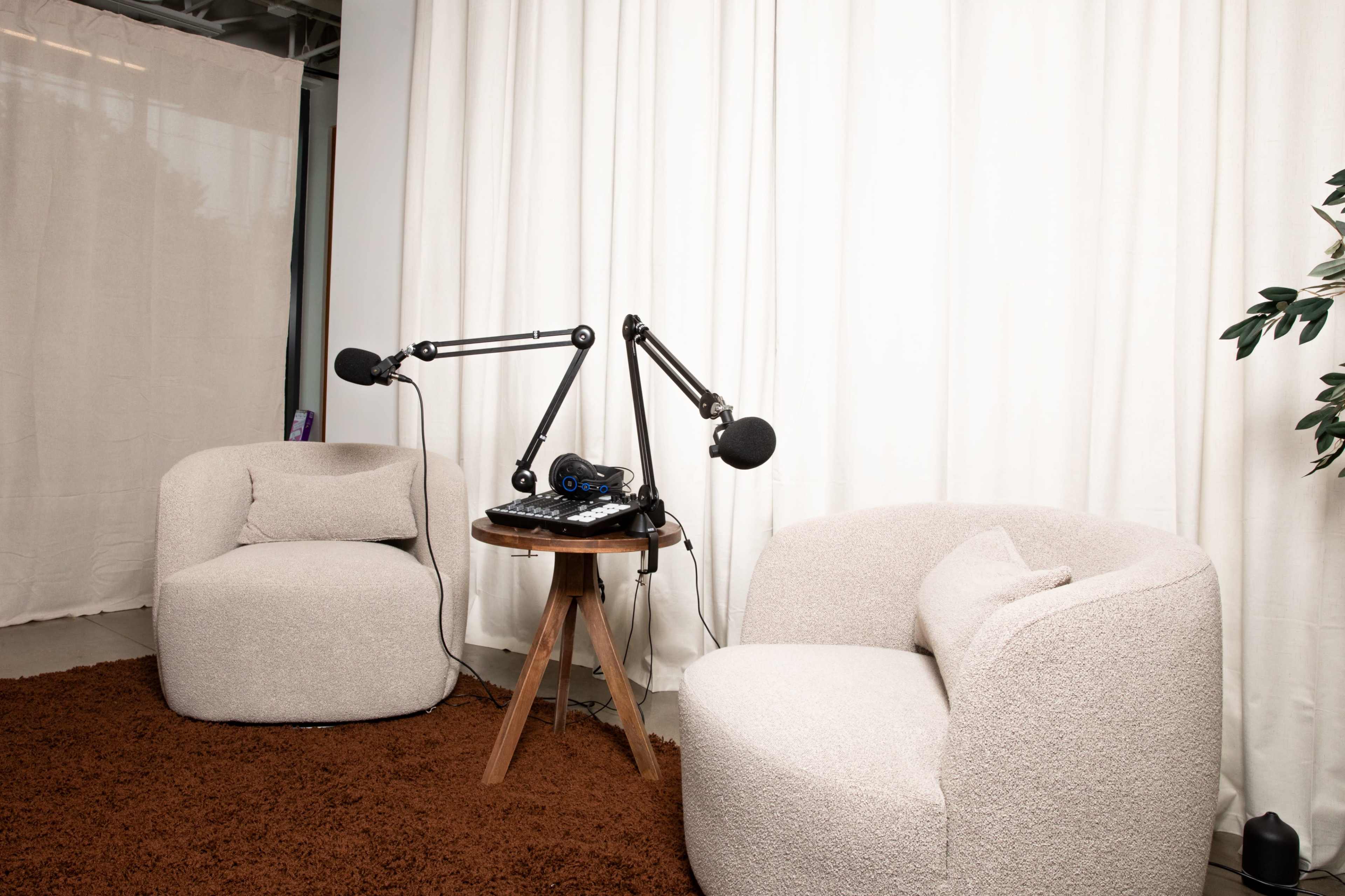 The image shows a cozy podcasting setup with two plush chairs, a small wooden table, and a microphone setup in front of a white curtain.