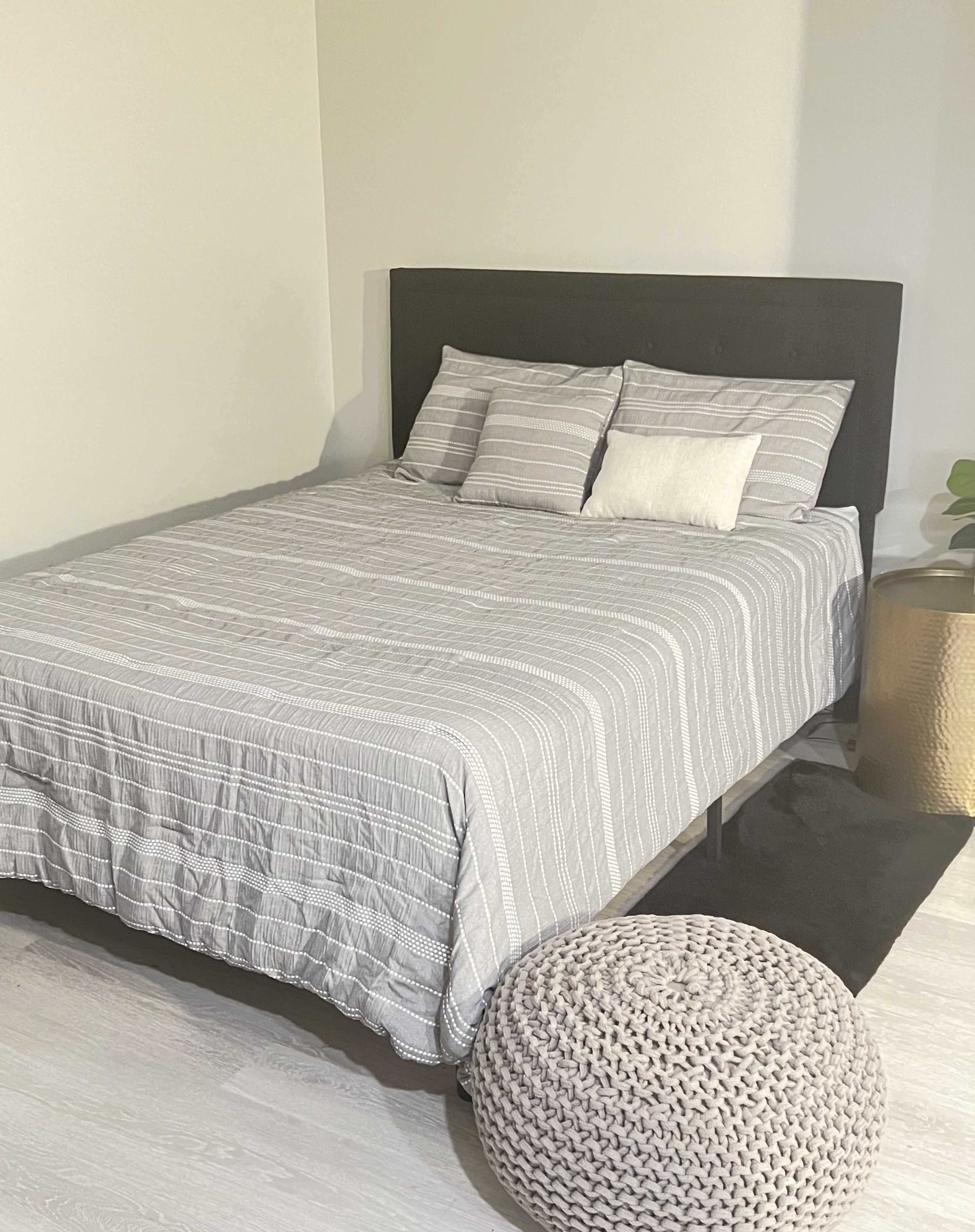 A simple bedroom with a bed covered in a gray striped quilt, two pillows, a small decorative pillow, a textured round pouf, and a potted plant beside a gold-colored side table.