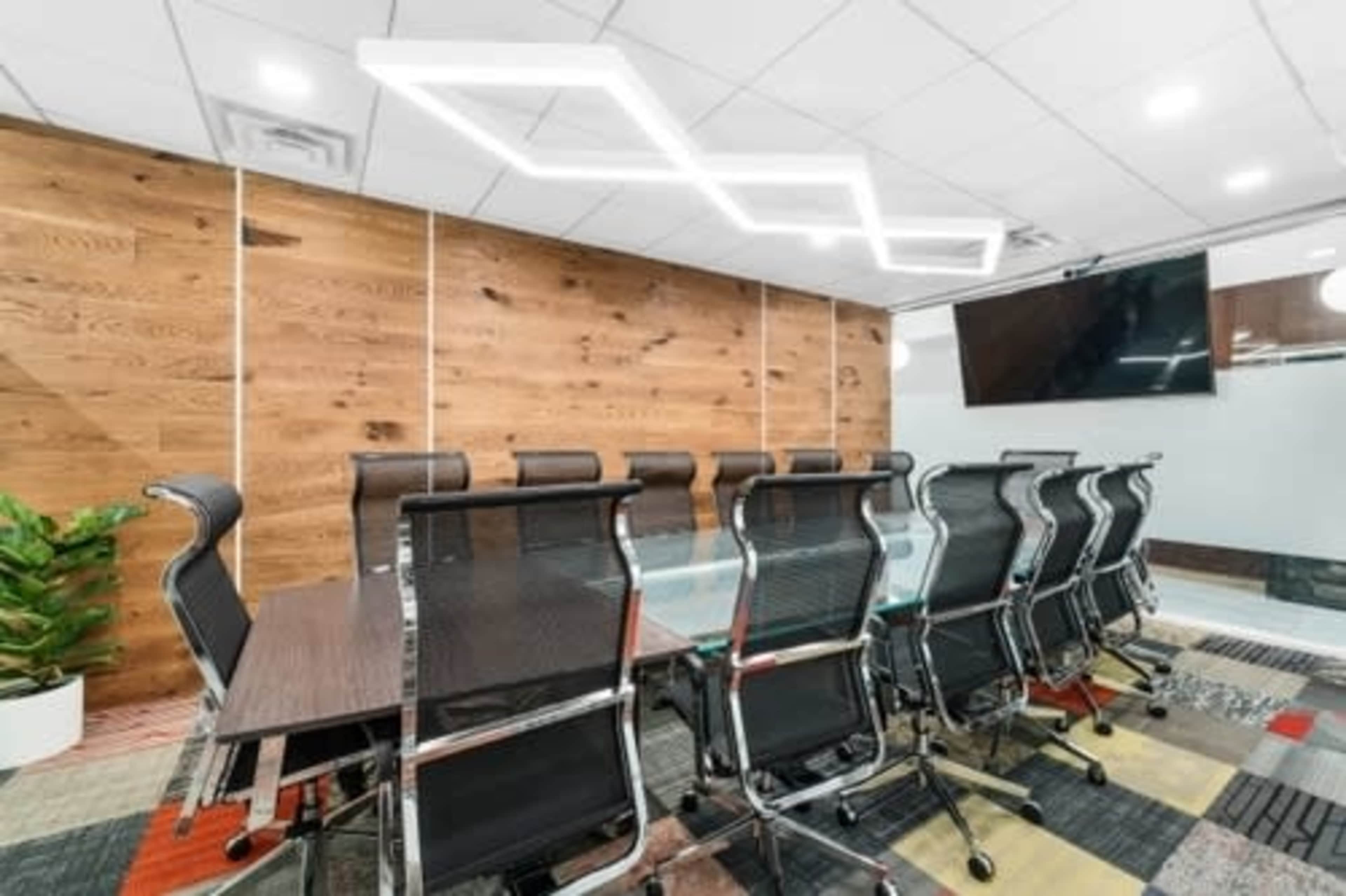 The image shows a modern conference room with a long table surrounded by sleek black chairs, a wood-paneled wall, and a geometric light fixture above.