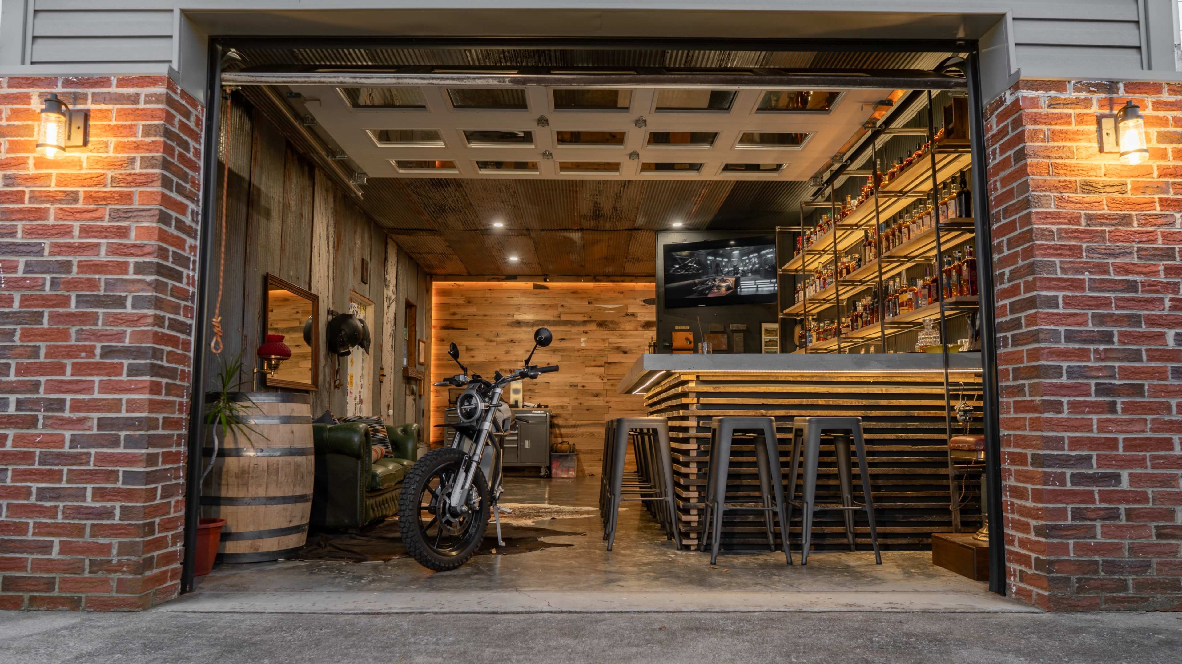 A motorcycle is parked inside a garage with a wooden bar and shelves in the background, and the garage door is open.