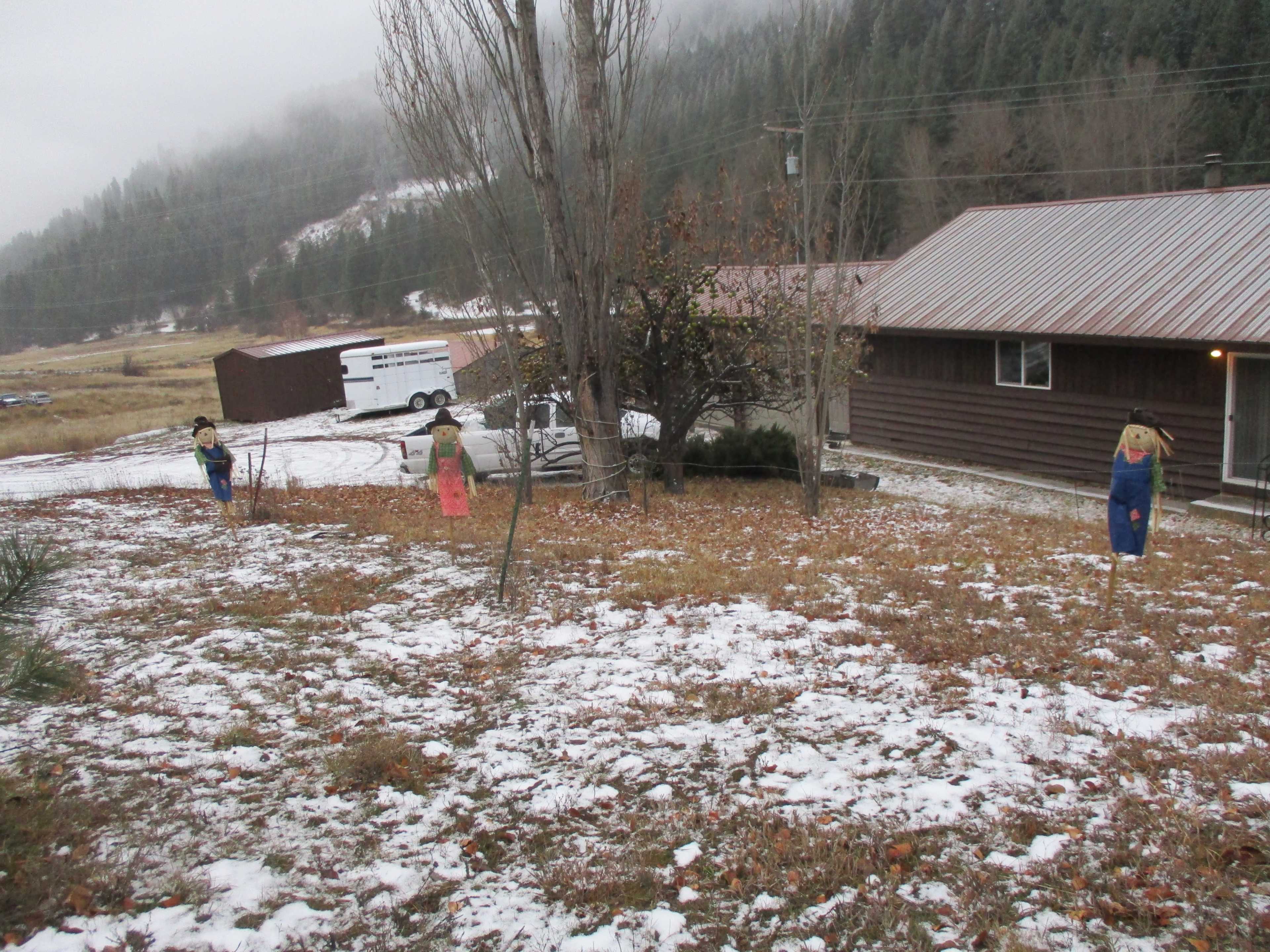 Three scarecrows stand in a yard covered with fallen leaves and patches of snow, set against a background of mountains and a wooden house.