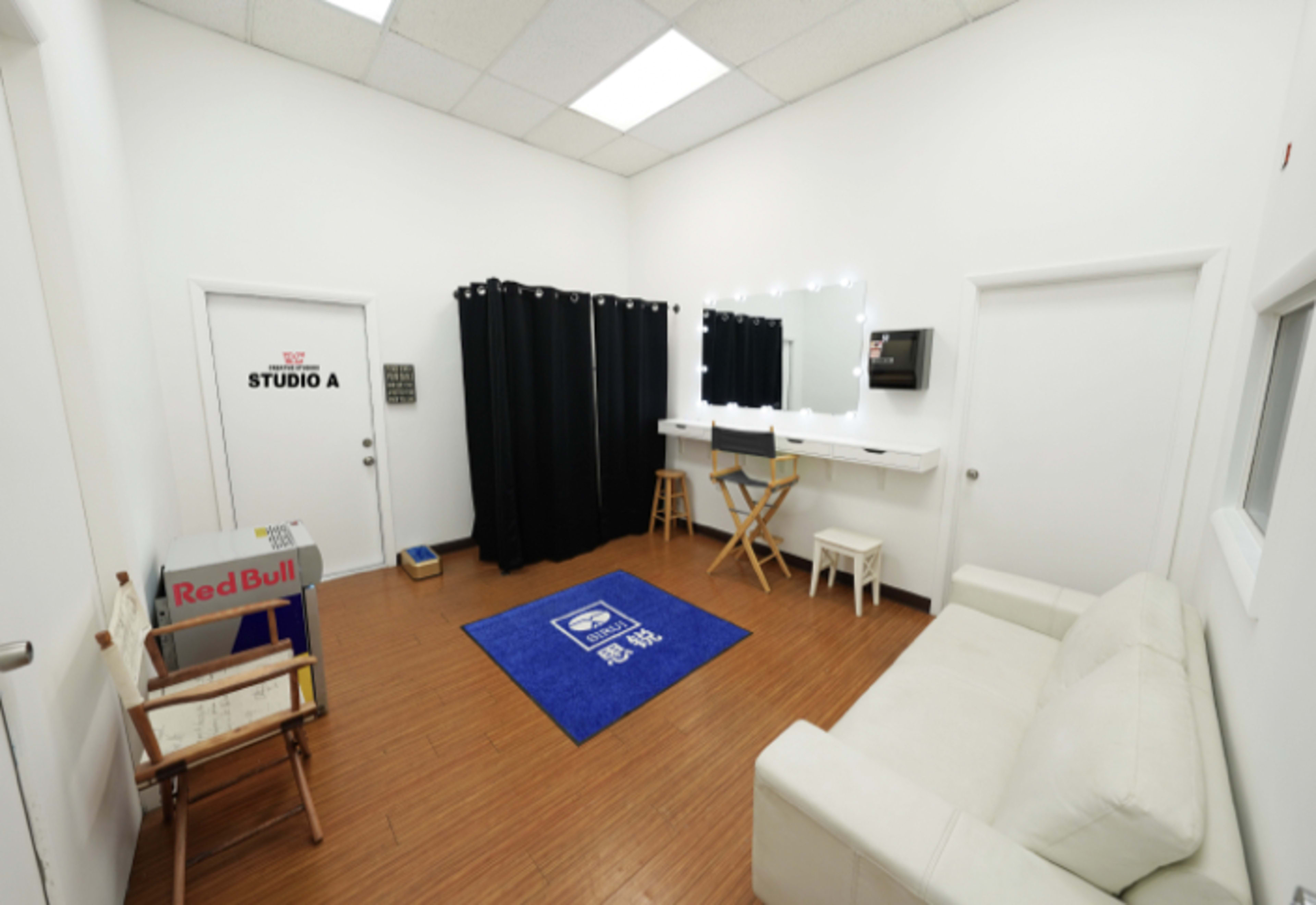 The image shows a small, modern studio room with a white sofa, a blue rug, a mirror with lights, and a curtain, along with a door labeled "STUDIO A."