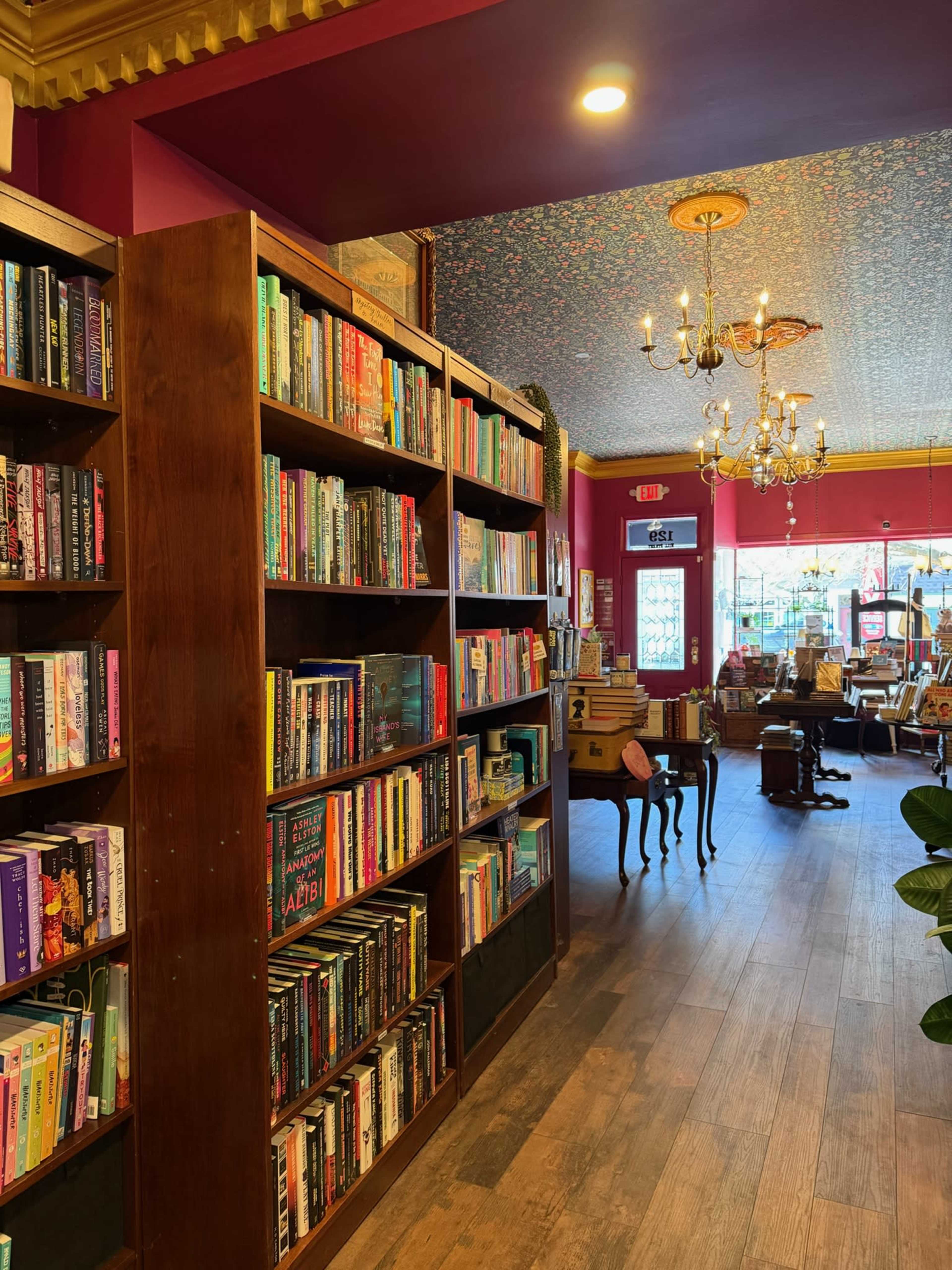 The image shows a bookstore interior with wooden bookshelves filled with colorful books, a chandelier hanging from the ceiling, and a cozy reading area in the background.