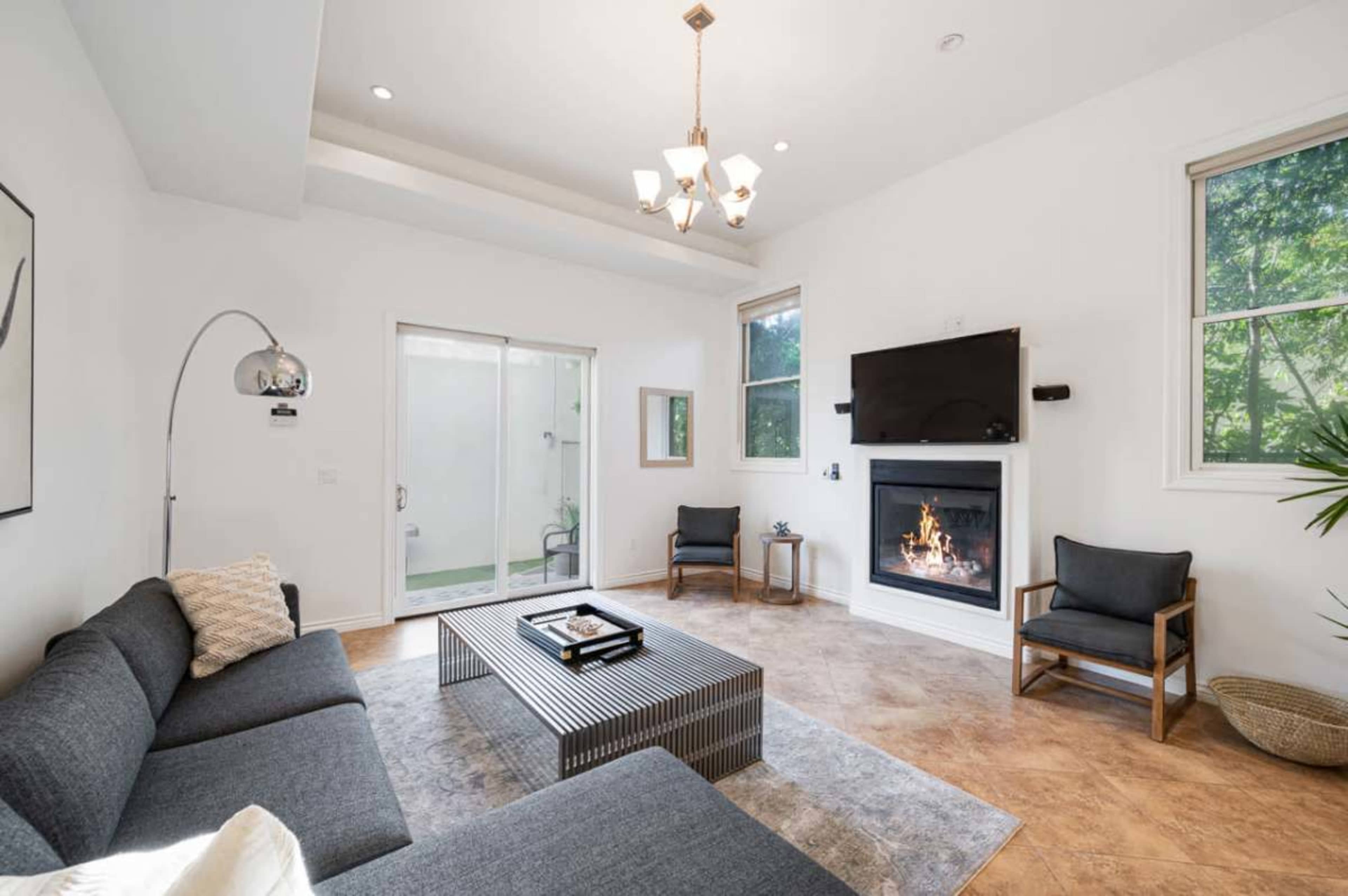 The image shows a bright living room featuring a modern fireplace, a coffee table, a sectional sofa, and two armchairs, with large windows allowing natural light to enter.
